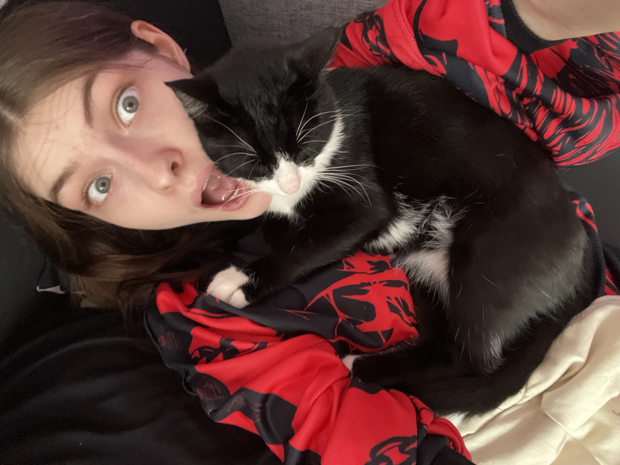 A woman lies on her back with a black and white cat resting on her chest. The woman is light-skinned with brown hair and wide, light-colored eyes. Her mouth is open. She is wearing a red and black patterned garment. The cat is mostly black with white markings on its chest, paws, and nose. The background consists of dark fabric, possibly a couch or bed. The location is indeterminable.