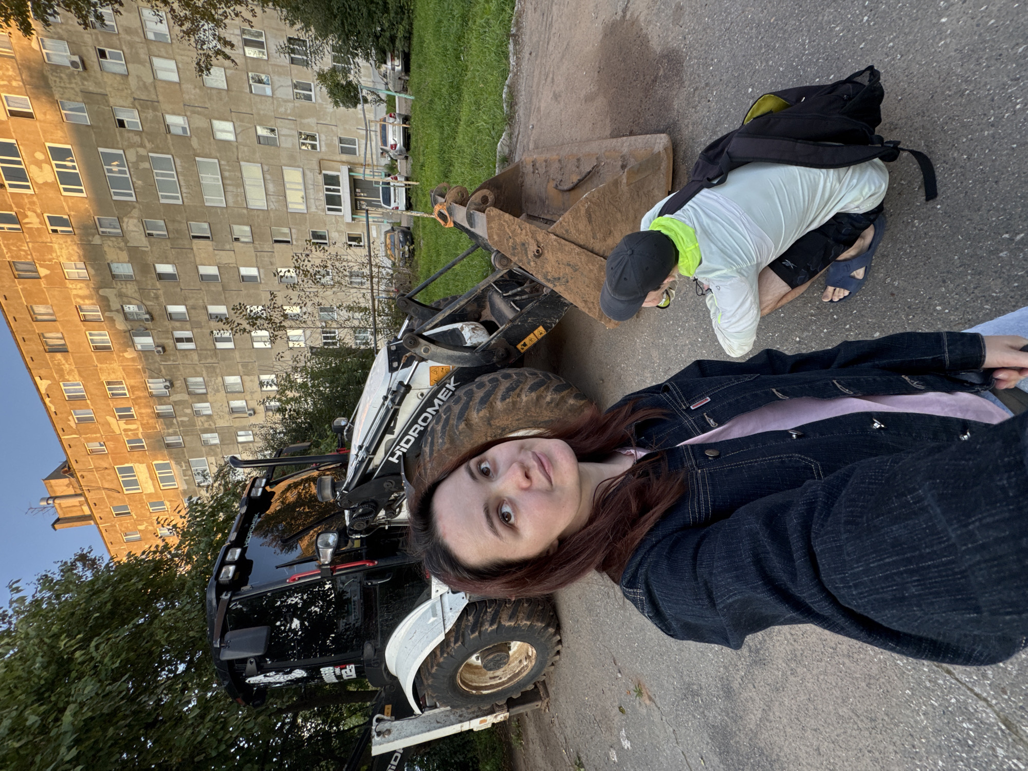 In Izhevsk, Russia, a young woman with dark brown hair, wearing a denim jacket over a pink t-shirt, takes a selfie in front of a white and black Hydromek backhoe loader. The heavy machinery is parked on an asphalt surface, with a large, dirt-covered bucket prominent in the foreground. To the right, a person wearing a black baseball cap, a light green hooded jacket over a white long-sleeved shirt, black shorts, and blue flip-flops is squatting down, facing away from the camera. They have a black backpack with a yellow interior slung over their shoulders. Behind the backhoe loader and the people is a tall, aged apartment building with a weathered facade and numerous windows, characteristic of Soviet-era architecture. The building is bathed in the warm light of what appears to be late afternoon or early evening sun. Green trees and a patch of grass are visible to the left of the building, and several cars are parked in the background, suggesting a residential urban environment. The overall scene captures a moment of casual activity in a typical Izhevsk urban setting, with construction equipment present.