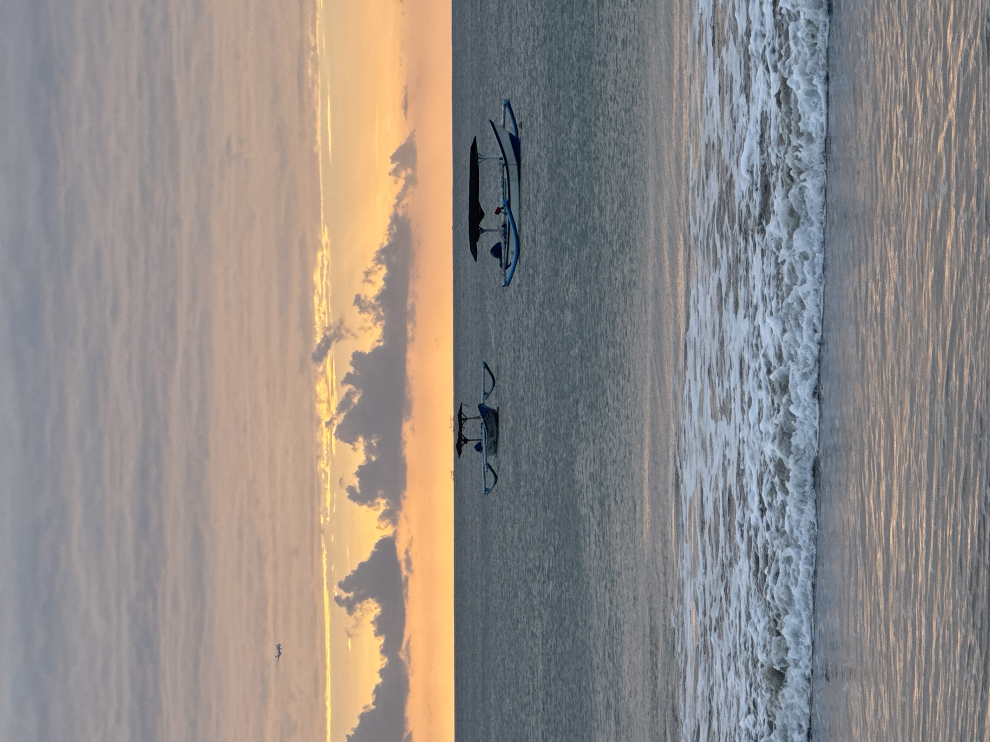 The image depicts a serene seascape at Kuta Selatan, Indonesia, during a sunrise or sunset. Two traditional Balinese outrigger boats are anchored on calm, greyish-blue waters. Each boat features a dark canopy over its center. In the distance, a small airplane is visible against a sky painted with soft orange and yellow hues, layered with undulating grey clouds. The foreground shows gentle waves breaking onto a sandy shore, with the water reflecting the warm colors of the sky. The scene conveys tranquility and the beauty of the coastal environment.
