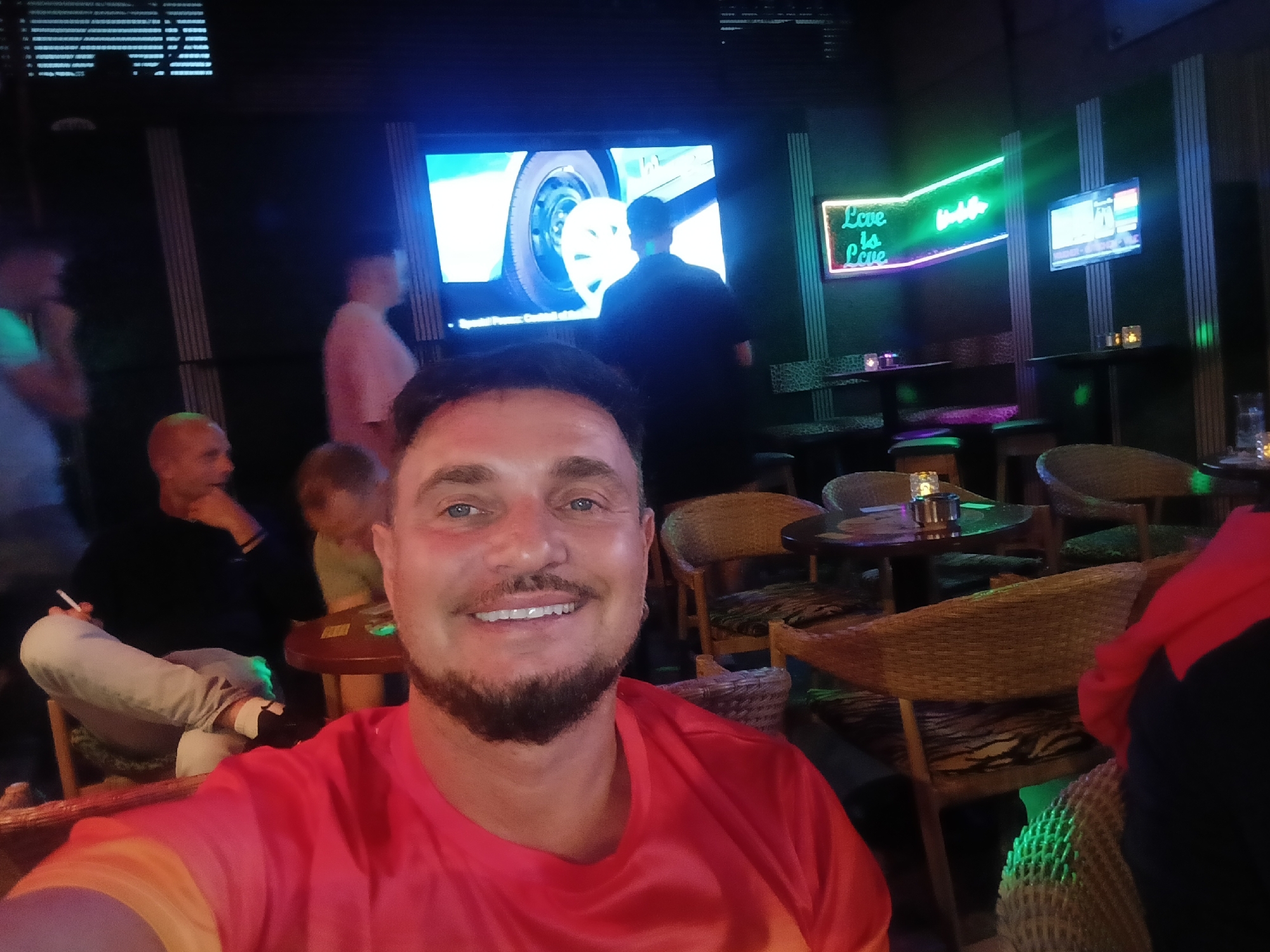 The photograph was taken in an indoor bar or lounge setting in San Bartolomé de Tirajana, Spain. The main subject is a man wearing a red t-shirt, smiling directly at the camera. He has short dark hair, blue eyes, and a beard. In the background, there are other people seated at tables, with one person holding a cigarette. The interior is dimly lit with a dark color palette. There's a TV screen showing an image of a car tire and a man in dark clothing. A neon sign that reads "Love is Love" in bright green light is visible on the wall behind the seated patrons. A smaller TV screen also adorns the wall in the background. The furniture appears to be wicker or rattan-style chairs and small round tables with lit candles.