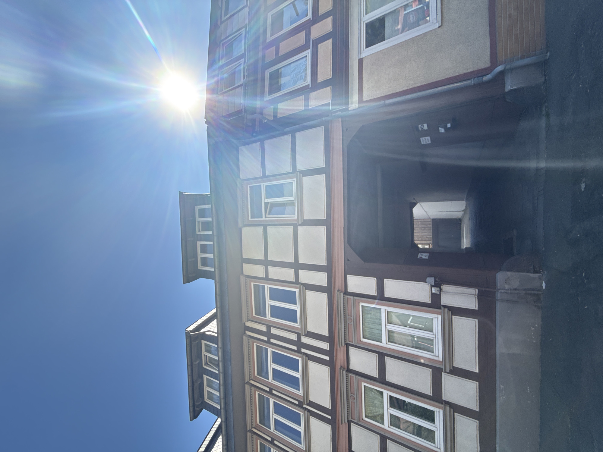 This low-angle, full-frame vertical photograph captures the facade of a historic timber-framed building in Bad Lauterberg im Harz, Germany. The building features a repeating pattern of dark brown timbers against a light beige infill, with several large, white-framed windows. Some windows have white curtains visible. The upper levels of the building have dormer windows.
A bright sun, positioned in the upper right quadrant of the image, creates a lens flare with distinct rays emanating outward and causing overexposure in its immediate vicinity. The sky above is a clear, vibrant blue.
The lower portion of the building reveals an arched entryway or passage leading into a darker interior. The ground in front of the building is paved with asphalt, showing some wear. A downspout runs vertically along the right side of the building. The architecture suggests a well-preserved historical structure within the town. No people are visible in the image.