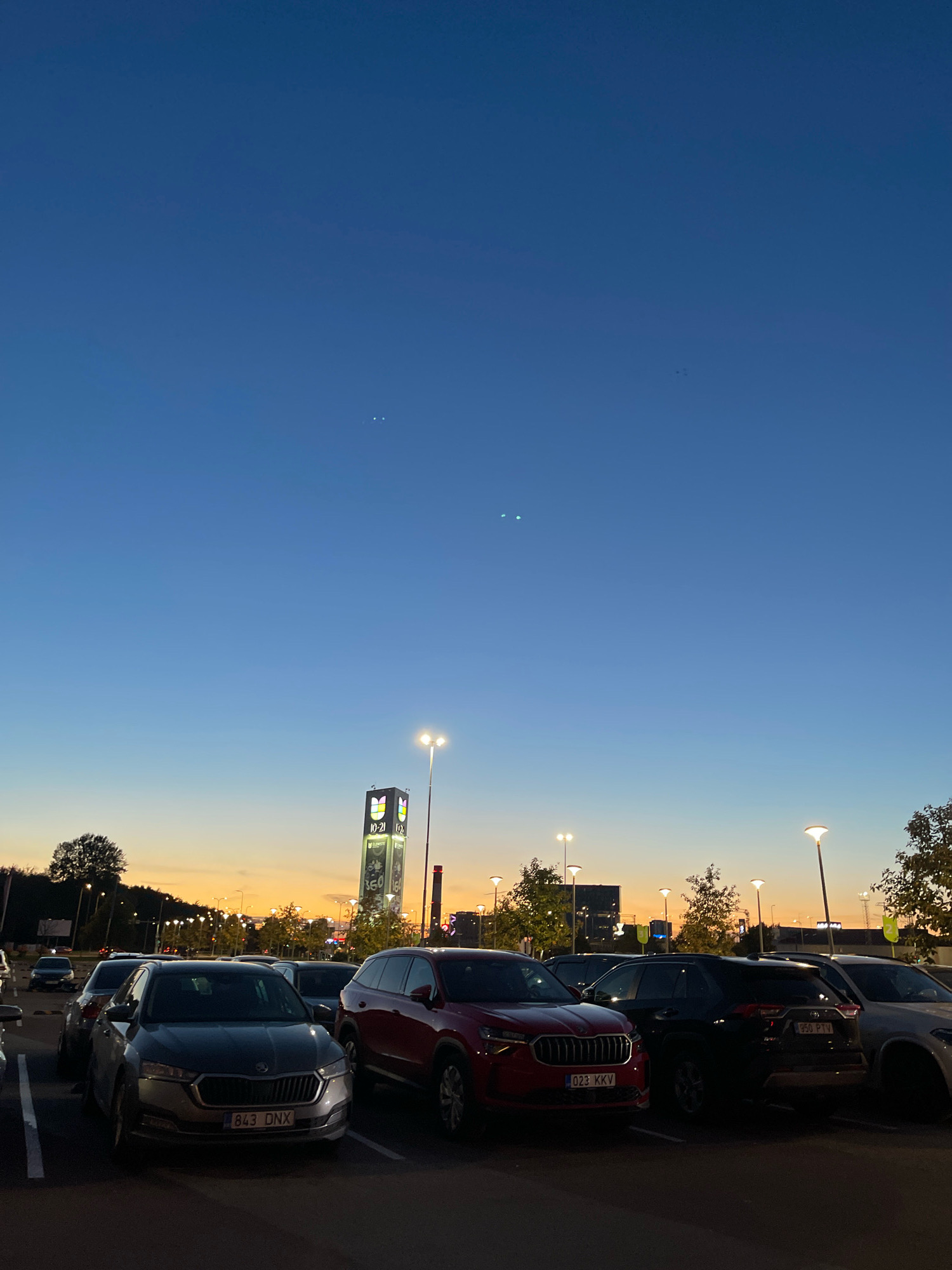 The image depicts a parking lot in Tallinn, Estonia, at dusk. In the foreground, several cars are parked. One prominent car in the center has the license plate "843 DNX." To its right, a red car with license plate "023 KKV" is visible, followed by a black SUV with license plate "950 PTV." In the background, a tall tower with illuminated logos, likely a commercial building or landmark, stands against the twilight sky. Streetlights are scattered throughout the parking area and along the horizon, illuminating the scene. The sky transitions from a deep blue at the top to orange and yellow hues near the horizon, indicating the setting sun. Two distinct pairs of bright, small lights are visible in the upper portion of the sky.