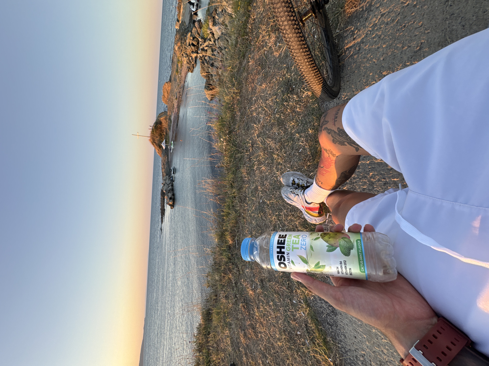 The image shows a first-person perspective of a person sitting on a grassy area overlooking the sea at sunset. In the foreground, the person's hand holds a bottle of "OSHEE VITAMIN GREEN TEA ZERO" with a "CACTUS-MINT FLAVOUR" label. The person is wearing white shorts, white sneakers with colorful accents, and white socks. A bicycle wheel and part of the frame are visible to the right. The background features the calm sea, a rocky coastline, and a small island with a cross on top. The sky is clear with a gradient of orange and blue hues, indicating sunset. This scene is likely located near the Church of St. George in Lozenets, Bulgaria, given the presence of the cross on the island and the coastal setting.
