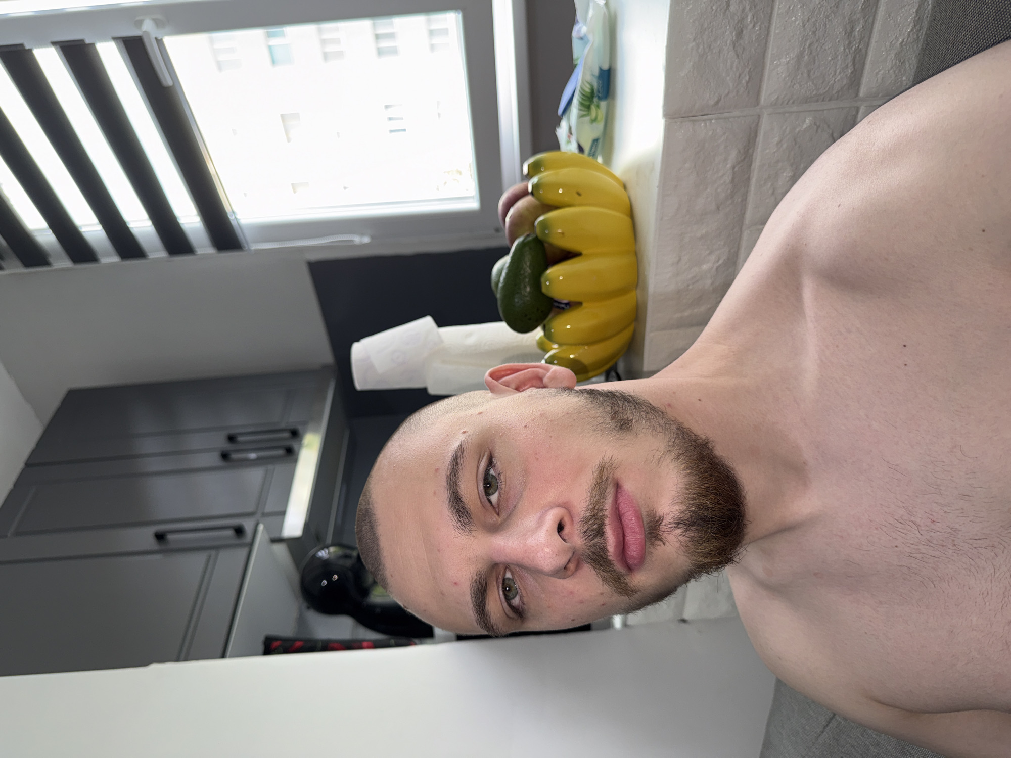 A shirtless man with a buzz cut and a beard is positioned in the foreground. Behind him, a kitchen setting includes gray cabinets and a window with blinds. A yellow banana-shaped container holds fruit including avocados and apples, sitting on a tiled surface next to the window. Further in the background, exterior buildings can be partially seen through the window. The location is Stara Zagora, Bulgaria.