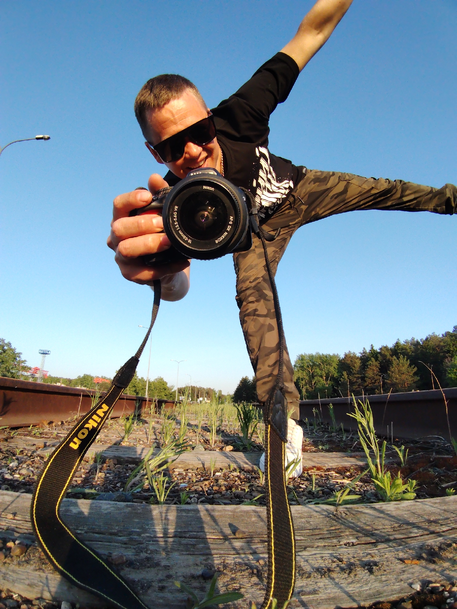 A man wearing sunglasses, a black t-shirt, and camouflage pants poses dynamically with a Nikon camera on a railway track. He stands with one leg extended in the air, arms spread, and holds the camera pointed toward the viewer. The camera has a black Nikon strap with yellow lettering, extending down to the wooden rail ties. The background includes more railway tracks with vegetation growing between them. In the distance are trees, street lights, and a structure resembling a tower. The sky above is clear and blue. The image appears to be taken at a low angle with a wide-angle lens, giving a distorted perspective. The location is in the city of Chaikovsky, Russia.