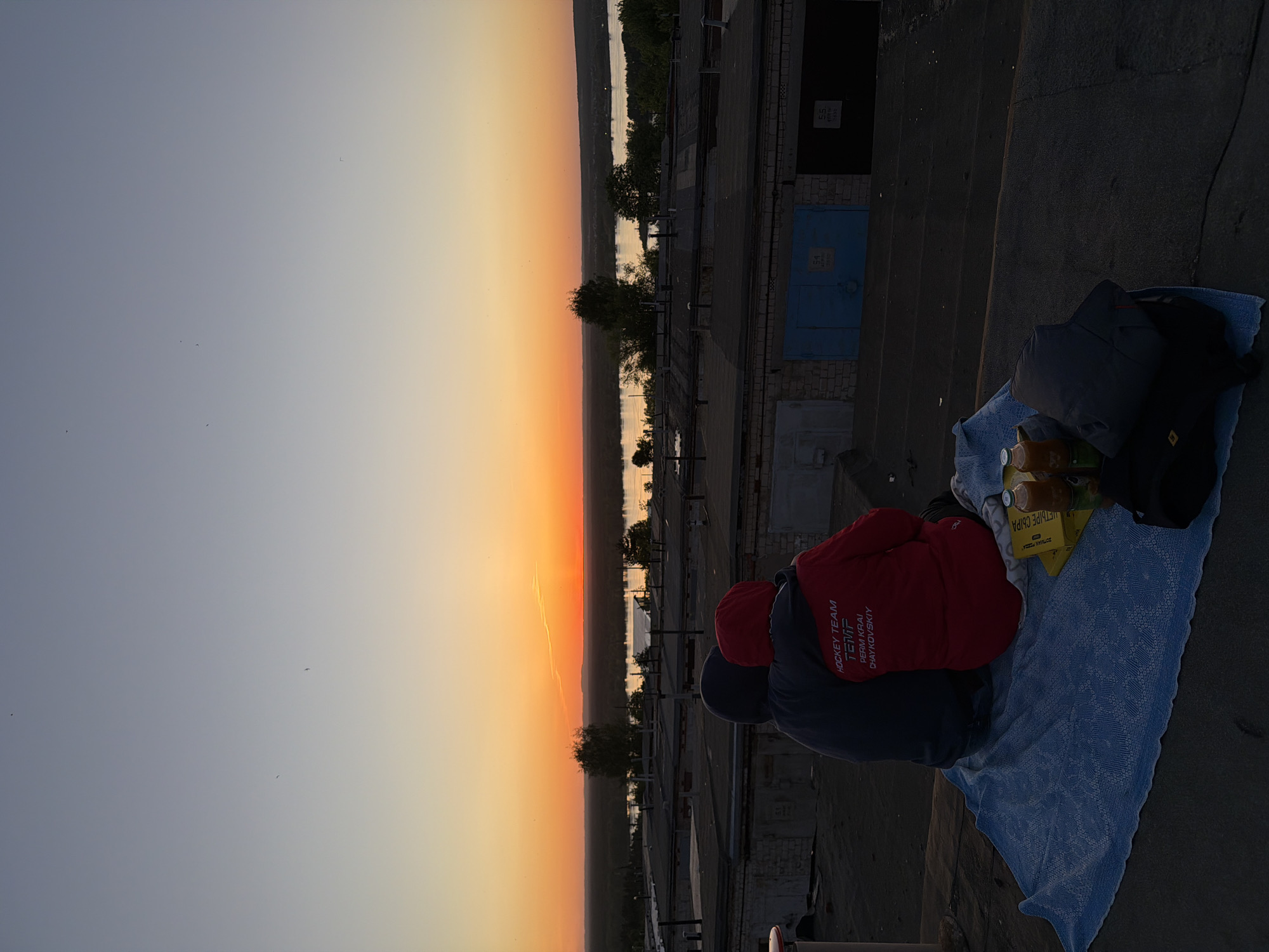 Two people are sitting on a blue blanket on a rooftop, facing away from the camera and toward a sunset. They appear to be enjoying the view with bottles and a yellow package of juice or beverage next to them. One person is wearing a red jacket with text on the back. This appears to be in Chaikovsky, Russia. The text on the back says "HOCKEY TEAM TEMP PERM KRAI CHAYKOVSKIY"