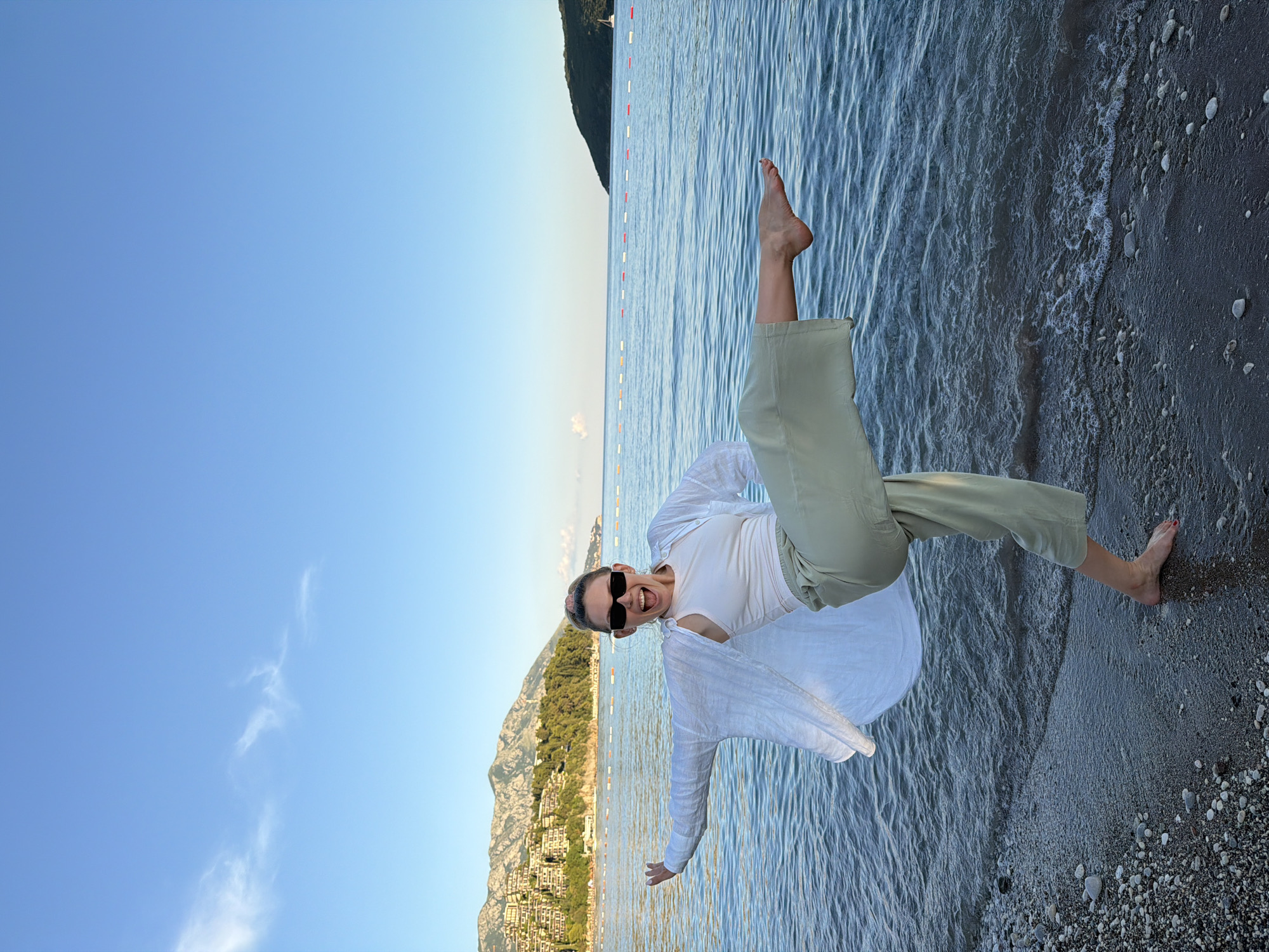 A woman stands on a pebble beach in Budva, Montenegro, with the Adriatic Sea behind her. She is wearing a white tank top, a white linen shirt, and loose green pants. Her left leg is extended forward, and her right leg is bent. Her arms are outstretched, and she has a wide, excited expression. The water is a deep blue, and there are mountains in the background. A line of buoys marks the swimming area. The sky is clear and blue, with a few wispy clouds. The sun appears to be setting or rising, casting a warm glow on the scene.