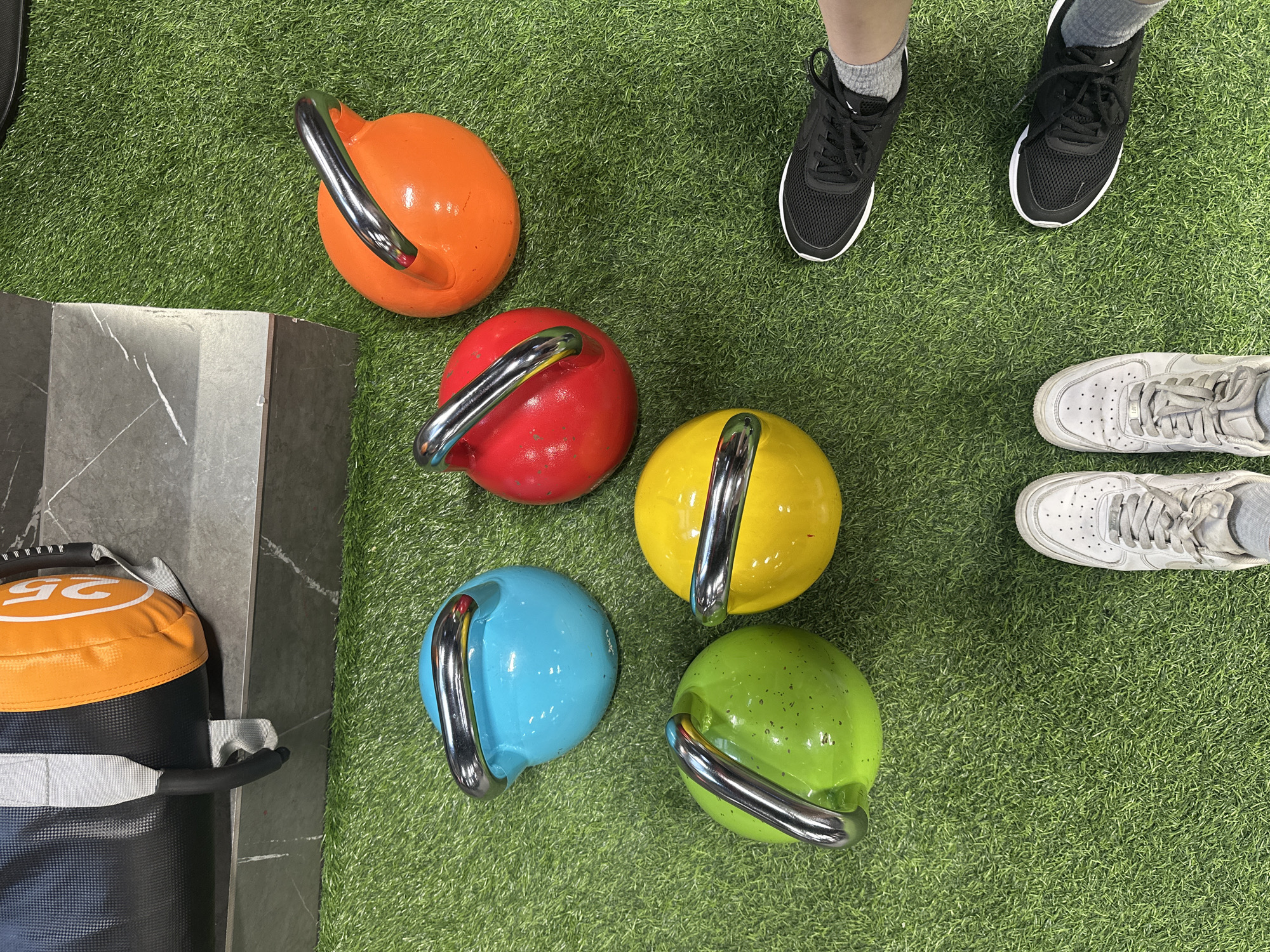 The image presents a gym or fitness area with an artificial green turf floor. Five brightly colored kettlebells (orange, red, yellow, blue, and green) are arranged in a cluster. A black and orange weight bag stands against a gray marble pillar. Two pairs of feet wearing athletic shoes (one black, one white) are also present, suggesting people are nearby. The location appears to be indoors in the city of Yerevan, Armenia.