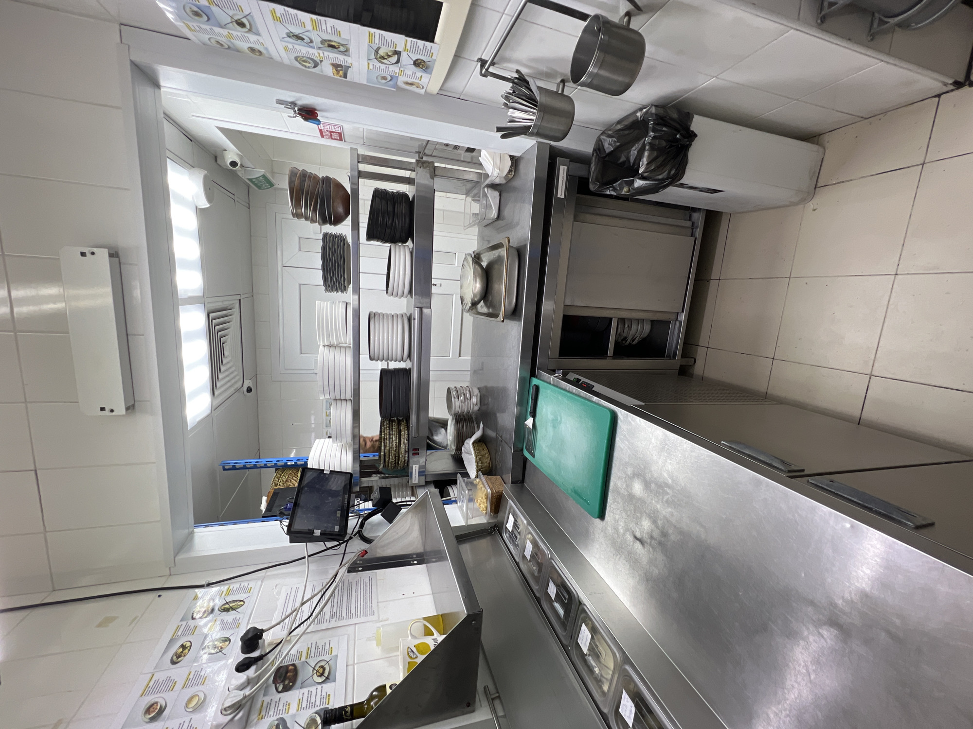 The image depicts a commercial kitchen in Plast Univska, Russia. Stainless steel countertops dominate the scene, featuring various kitchen implements and storage solutions. To the left, a row of transparent food containers sits within a refrigerated unit. A green cutting board with a knife is prominently placed on the counter. Shelving units display stacks of white and dark plates, along with bowls. A trash can with a black liner is positioned on the floor. A surveillance camera is visible near the ceiling, suggesting security measures. The walls are tiled with white squares. The overall environment appears organized and functional, typical of a professional kitchen.