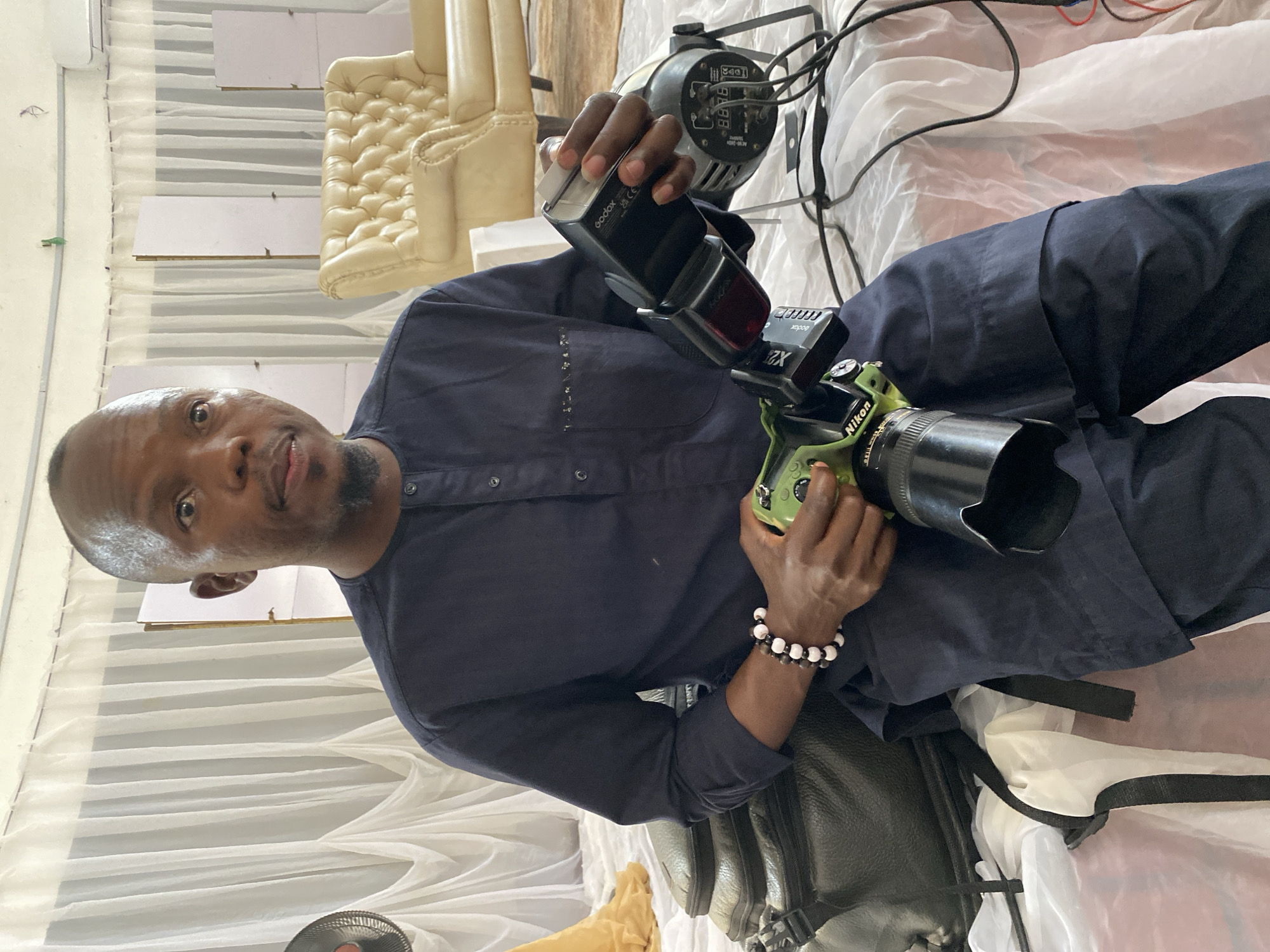 This media file captures a close-up, eye-level view of a man holding a camera and a flash unit, seemingly in a studio or indoor setting. The man is wearing a dark blue, collared shirt with several buttons. He has a short beard and is looking directly at the camera with a neutral expression. On his left wrist, he wears a beaded bracelet.
In his hands, he holds a green Nikon camera with a large black lens attached. The camera is angled slightly towards the viewer. Above the camera, he is holding a black flash unit, labeled "Godox".
The background consists of draped white fabric, creating a soft, diffused backdrop. To the left of the man, a portion of a tufted, cream-colored chair is visible. Behind the man and the fabric, a professional lighting equipment, possibly a continuous light or a studio strobe, is partially visible, with wires extending from it. There are also elements that suggest a studio environment, such as a white partition or backdrop.
The lighting appears to be artificial, consistent with an indoor photography setup. The overall impression is that the man is a photographer, possibly setting up for or in the middle of a photo shoot. The image is oriented vertically, with the subjects appearing sideways. The location context provided is Bukuru, Nigeria, though no specific visual cues within the image directly confirm this. No discernible weather or time of day can be determined from the image itself. Visible text includes "Godox" and "Nikon".