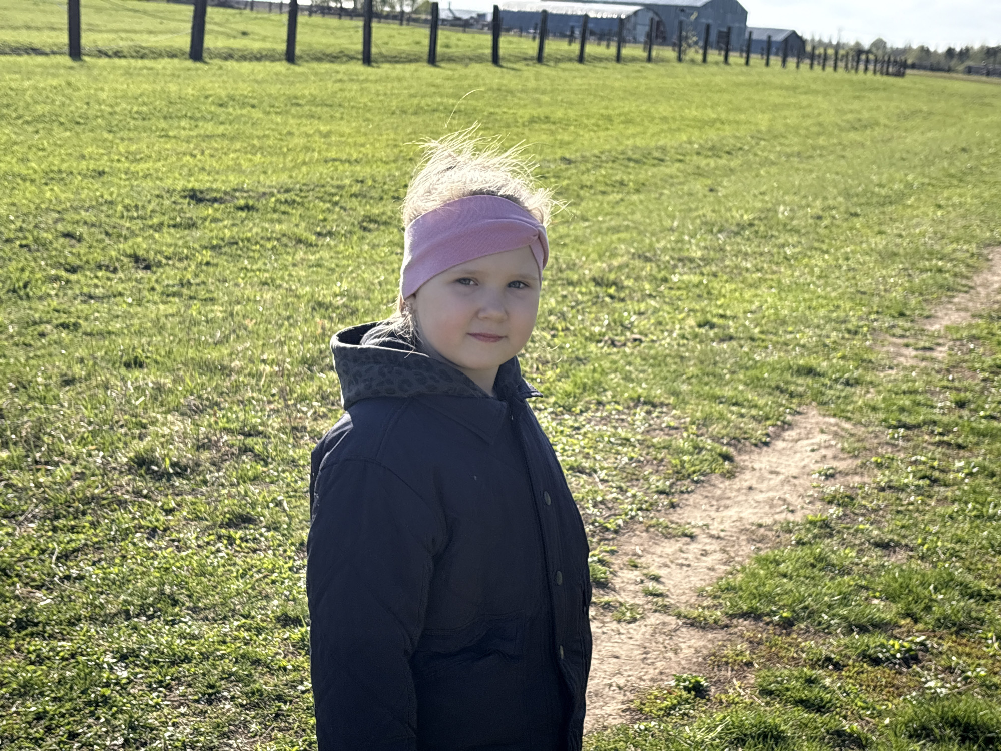 A young girl stands on a dirt path in a grassy field. She wears a pink headband and a dark blue jacket with a patterned hood. A fence line runs in the background, behind which are farm buildings. The location is most likely Азинский, Russia.