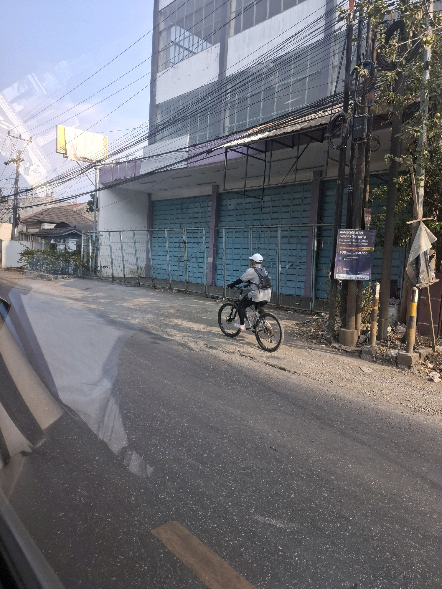 A person wearing a white cap, grey t-shirt, and black pants rides a silver bicycle down a paved road in Karangtengah, Indonesia. The cyclist has a black backpack on. The road is lined with a metal fence and a building with teal roller doors. Overhead, a complex network of power lines crisscrosses the sky, strung from utility poles. A tall, modern building with reflective windows looms behind the roller doors. To the right of the road, a tree with green leaves stands beside a utility pole adorned with a banner advertising internet services. The ground on the right side of the road is rough and covered with debris. The scene is bathed in the warm light of day.