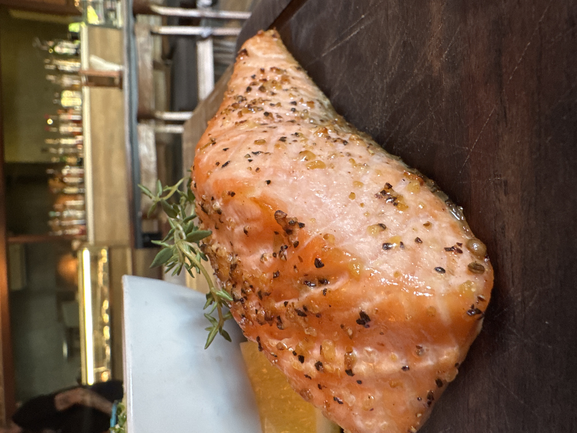 A close-up photograph shows a piece of cooked salmon resting on a wooden surface. The salmon is seasoned with herbs and spices. A sprig of fresh thyme is positioned beside the salmon. Part of a white plate or serving dish is visible beneath the salmon. In the background, out of focus, there is a shelving unit displaying bottles, suggesting a bar or restaurant setting. Based on the prompt, it is implied that this scene is located in Kuta, Indonesia, although this cannot be verified visually.