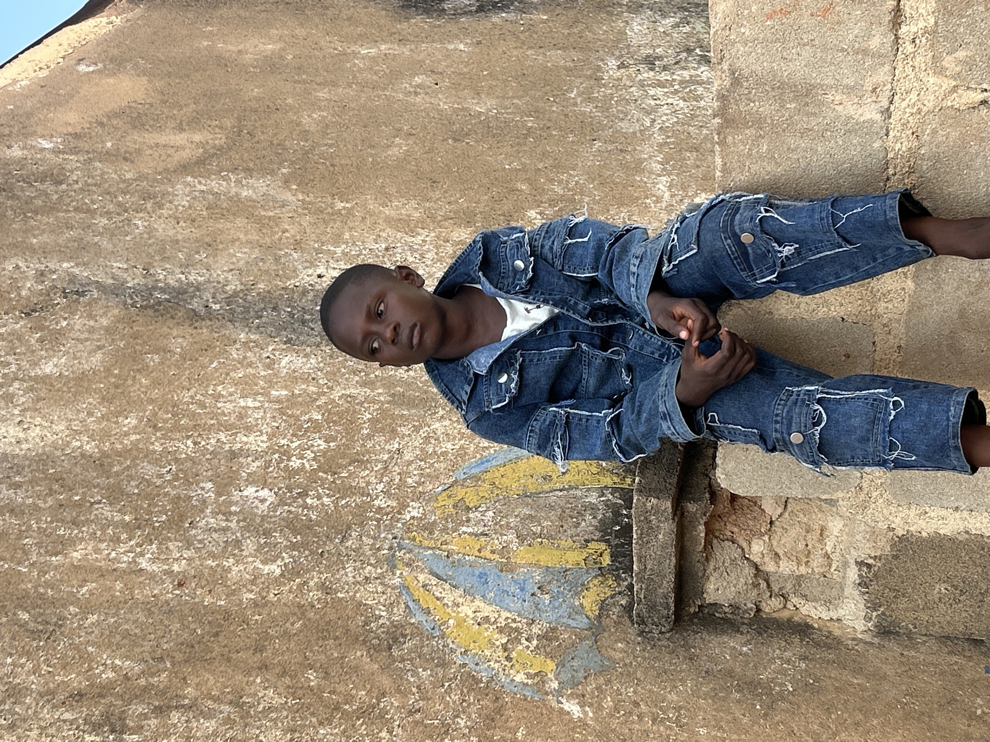 An outdoor, eye-level full-shot image captured in Jos, Nigeria, features a young dark-skinned boy, estimated to be between 6 and 10 years old, seated on a stone or concrete ledge against a textured, light-brown stucco or concrete wall. The boy wears a distressed, medium-blue denim jacket over a white t-shirt and matching distressed denim cargo pants, both exhibiting visible rips, fraying, and white stitching details. His short dark hair is neatly cut, and he gazes intently towards the left of the frame with a neutral, slightly pensive expression, his hands loosely clasped in front of him. To the left of the boy, painted onto the wall surface, is a faded, stylized depiction of an open umbrella with alternating yellow and light blue segments. The wall itself displays a rough, granular texture and variations in tone consistent with weathering and age. A narrow sliver of clear blue sky is visible in the upper right corner, indicating an outdoor, daylight setting. The overall lighting is bright and even, highlighting the textures of the wall and the boy's attire.