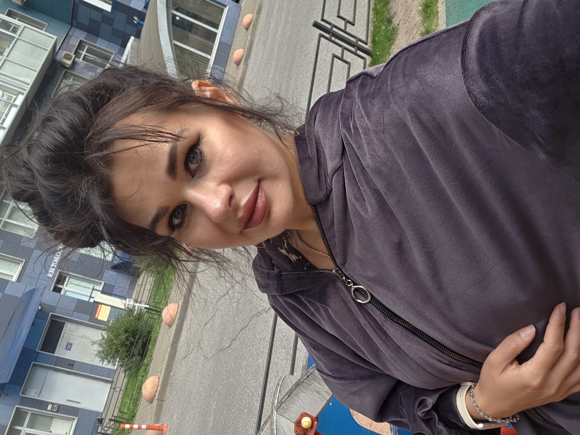 A woman with dark hair pulled into a messy bun poses for a selfie outdoors in Saint Petersburg, Russia. She wears a dark gray zip-up hoodie and a silver bracelet. In the background, a modern blue and gray building with multiple windows is visible, along with a sidewalk, orange and white traffic cones, and rounded concrete bollards. The environment suggests a residential or commercial area within the city.