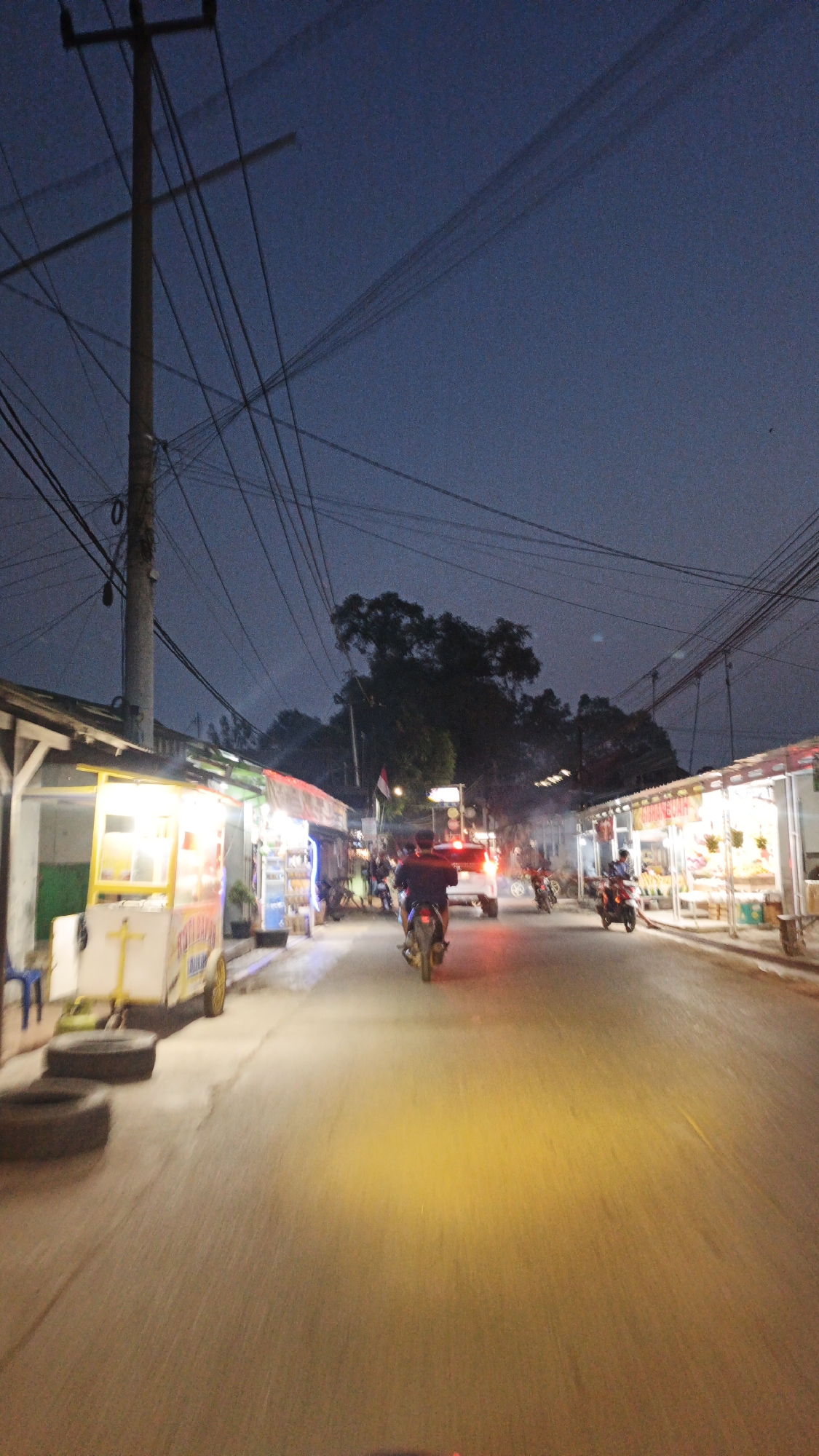 This is a photograph taken from a moving vehicle on a street in Muntur, Indonesia, during twilight. The scene depicts a street lined with small shops and food stalls, illuminated by artificial lighting. Several motorcycles are visible, with some being ridden by people. A white truck is also present on the road. Overhead, a complex network of electrical wires stretches across the dark blue sky, supported by a utility pole on the left side of the frame. The road surface is asphalt and reflects the lights from the shops and vehicle headlights, creating a warm, yellowish glow in the foreground. The overall atmosphere suggests a busy evening street scene in a semi-urban or rural Indonesian setting.
