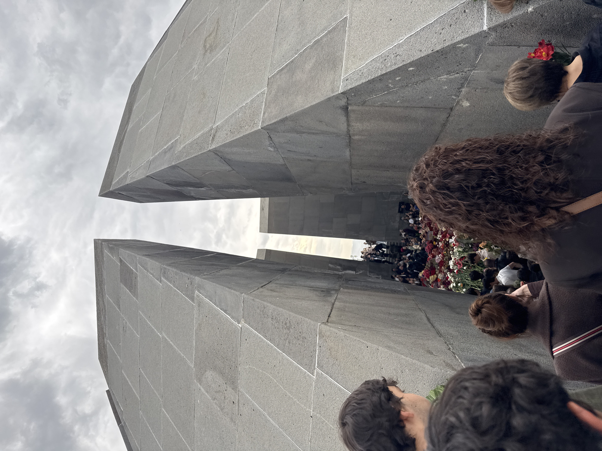 The image depicts a solemn gathering at a large memorial, likely the Tsitsernakaberd Armenian Genocide Memorial in Yerevan, Armenia, given the provided context. The perspective is a low-angle shot, looking upwards and forwards between two immense, converging grey structures made of large, rectangular stone blocks. These monumental pylons frame the central memorial area.
In the distance, between these imposing structures, a substantial crowd of people is gathered. Many are seen placing or observing a large collection of flowers, predominantly red and white, which form a vibrant mound at the base of the memorial. The foreground features the backs and sides of several individuals, their heads and shoulders visible, indicating they are part of the procession or attending the ceremony, all looking towards the memorial area.
The sky is overcast and grey, contributing to a somber and reflective atmosphere. The weather appears to be cloudy daytime. No visible text is present in the image. The scene conveys a sense of remembrance and commemoration, with the act of laying flowers being a central activity.
