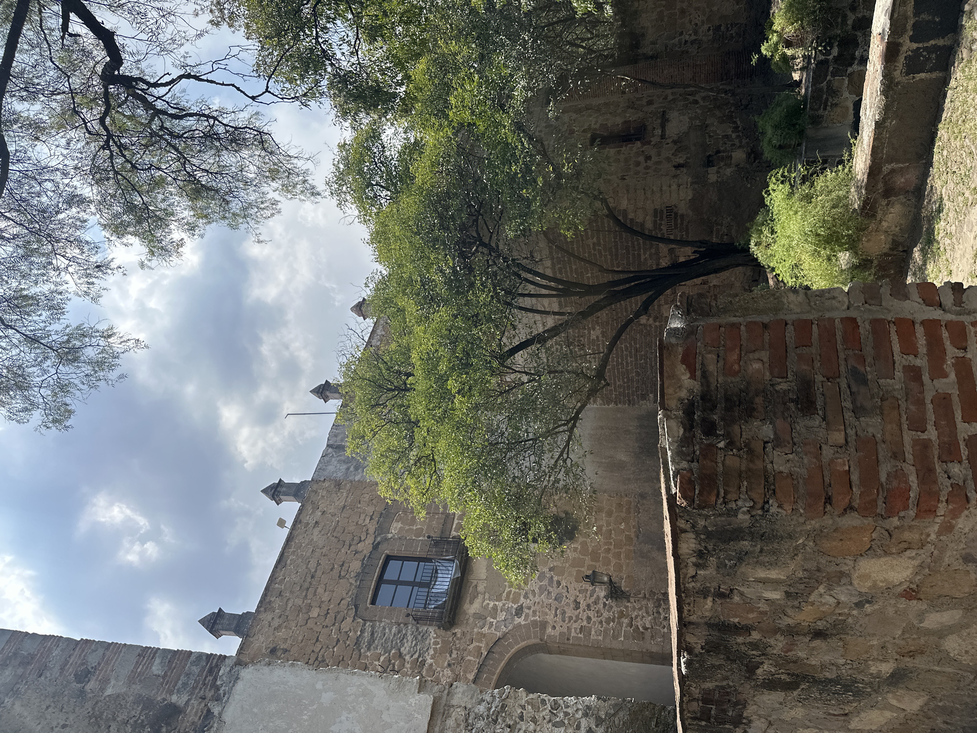 A low-angle shot captures the exterior of an old stone building with trees growing alongside it, possibly located in Tlalnepantla, Mexico. The building features a pointed roofline adorned with decorative spires. A dark-framed window is visible on the building's facade, which is constructed from uneven stones. A portion of the roof is covered by a tree with lush green leaves, partially obscuring the sky with white clouds overhead. The right side of the frame reveals a brick and stone wall, adding to the aged appearance of the setting. The scene conveys a sense of historical architecture blended with natural elements.