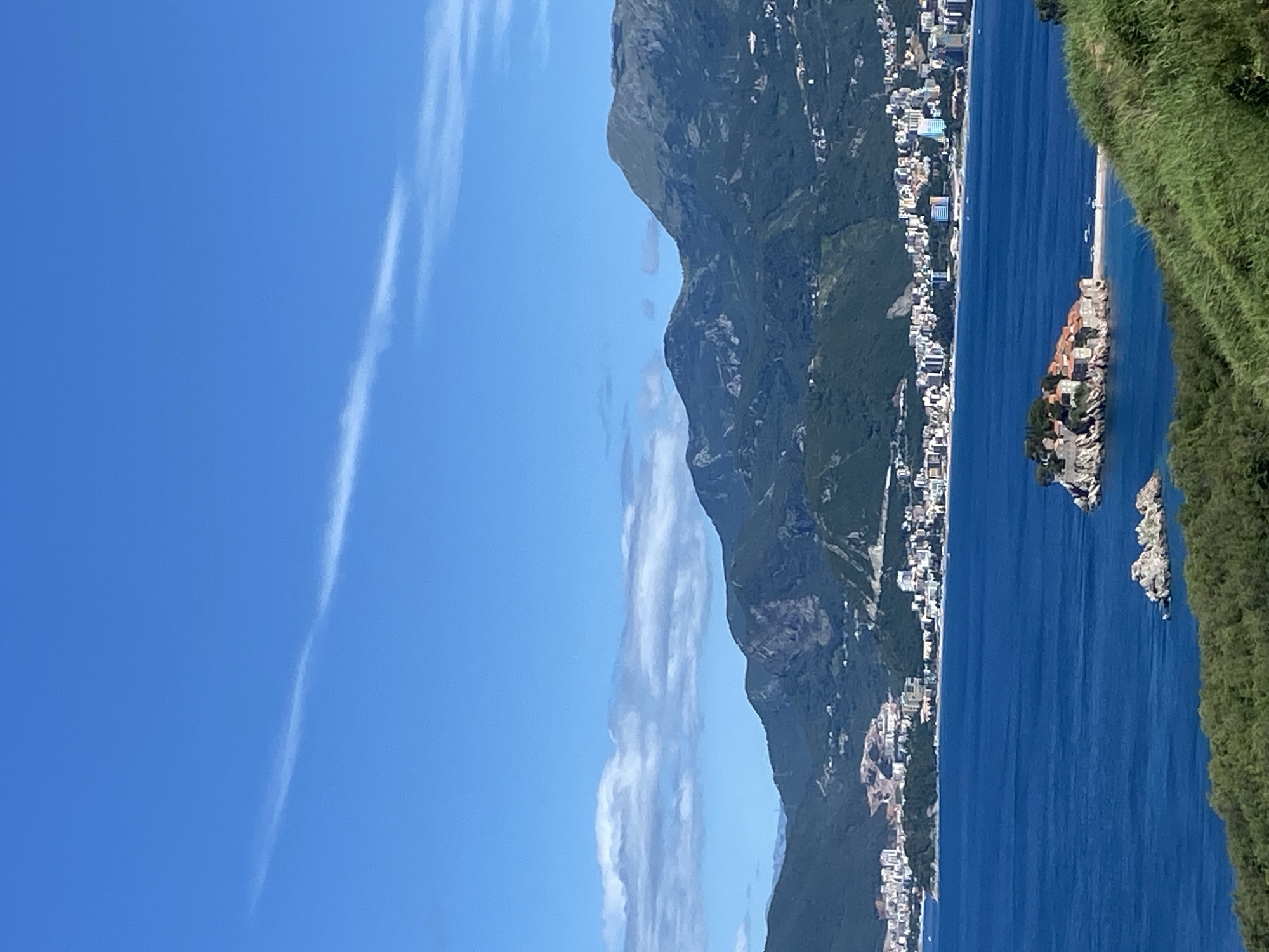 This is a landscape photograph taken on a clear, sunny day in Budva, Montenegro. The image captures a panoramic view of the Adriatic Sea, with a small island dotted with buildings in the foreground. The island appears to be connected to the mainland by a thin strip of land.
In the mid-ground, a coastline is lined with a dense concentration of buildings, suggesting a bustling city or town. These structures are a mix of modern and older architecture, with varying heights and colors. Behind the city, a range of lush green mountains rises steeply, their slopes covered in dense vegetation. The sky is a vibrant, clear blue with a few wispy white clouds streaking across it.
The foreground of the image shows a steep, grassy slope covered in verdant green foliage, implying an elevated vantage point for the photographer. The overall scene is one of natural beauty and coastal development.