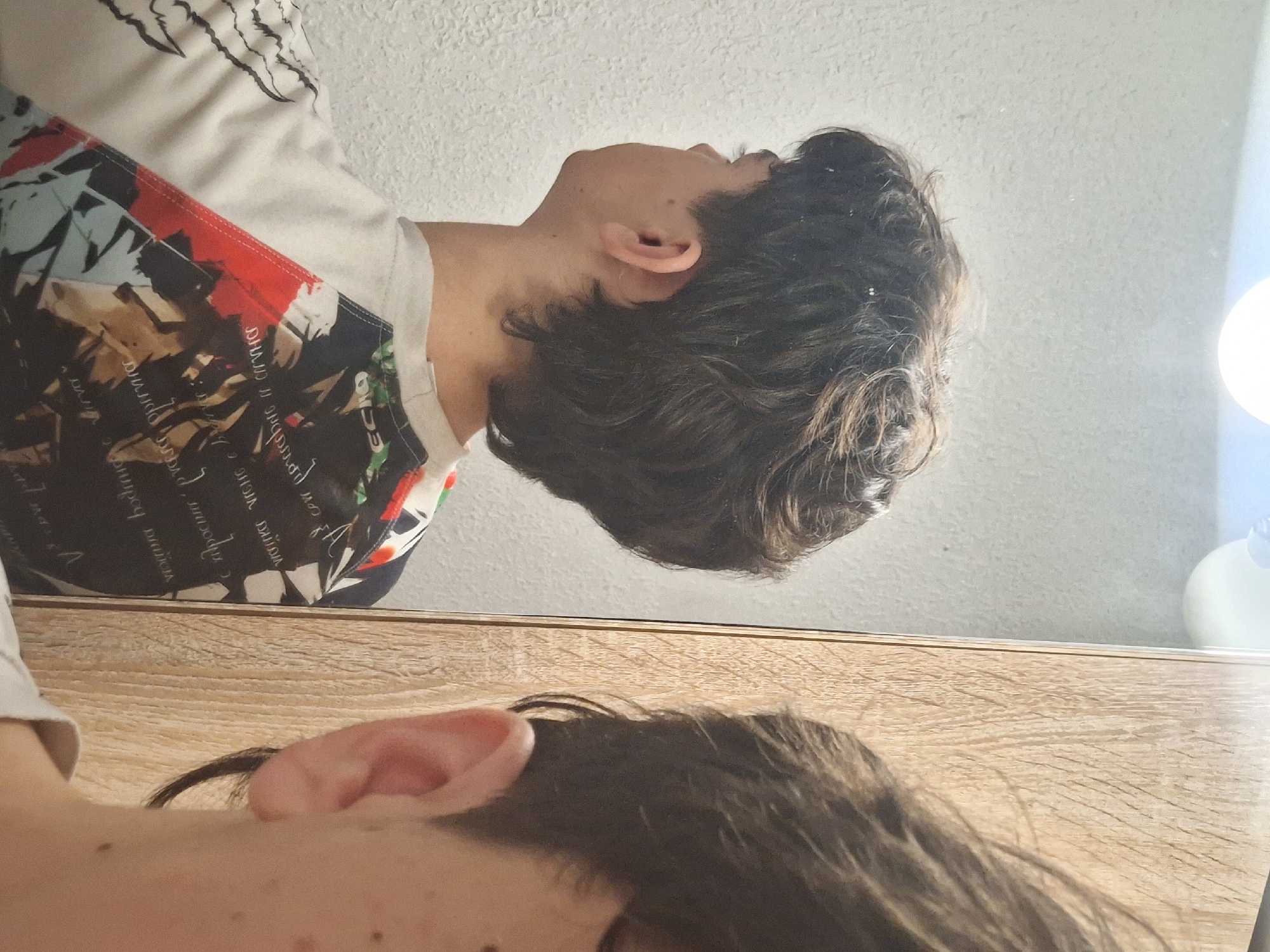 The image shows a person's reflection in a mirror. The person is wearing a t-shirt with a colorful, patterned design. The background appears to be a light-colored wall. The surface on which the mirror rests appears to be a wooden table. There is no location information visible in the image that would indicate the city of Burgas, Bulgaria.