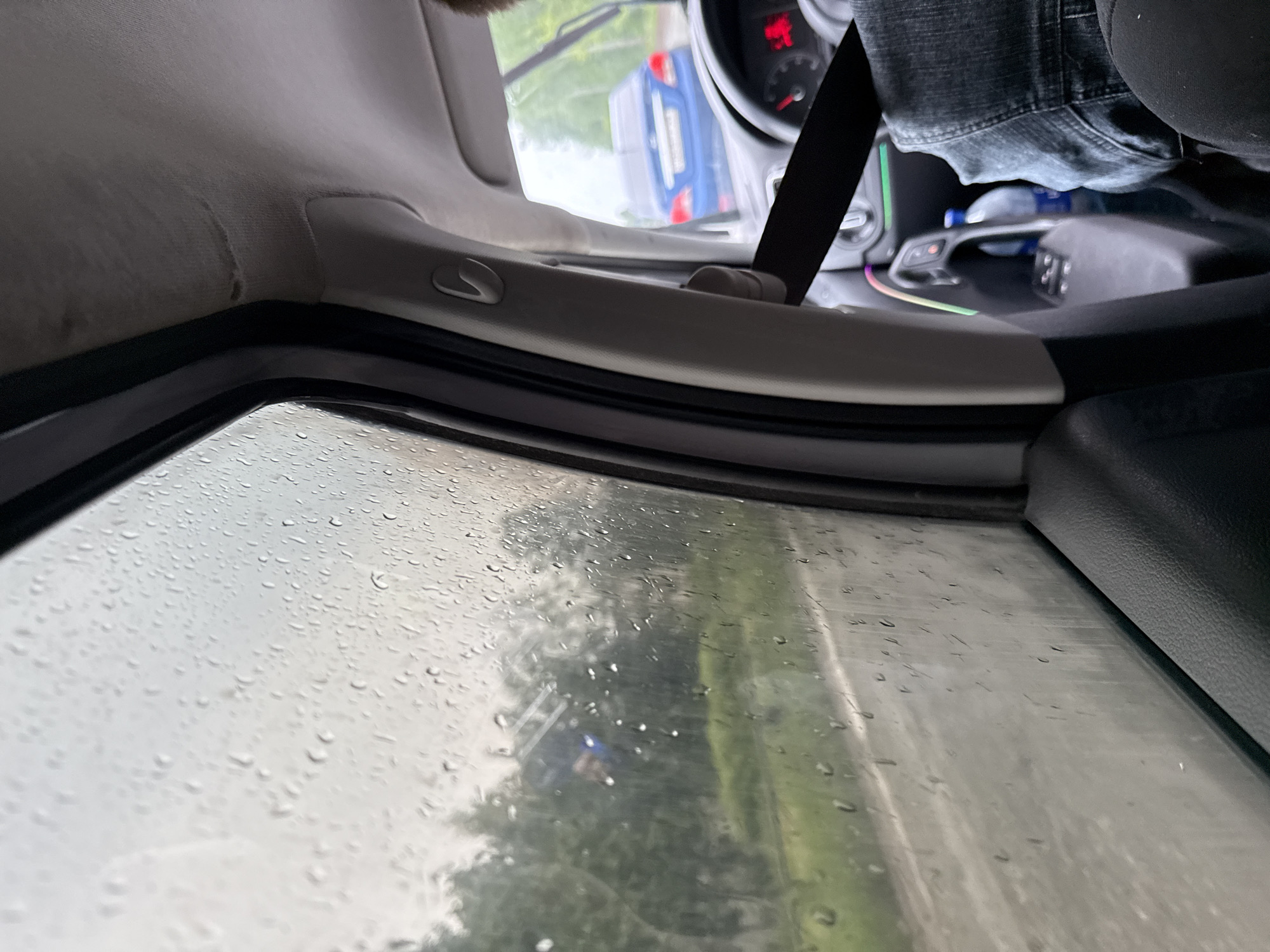 The image provides an interior view of a car, taken from a low angle looking towards the front-right side window during what appears to be a rainy day. The car's interior features a light-colored headliner with a noticeable dirty or water-stained patch, suggesting wear or a previous leak. Below this, grey plastic trim and a black seatbelt are visible, extending down towards the dashboard.
On the right side of the frame, the lower part of a person's legs, clad in dark blue jeans, is visible along with a black seat cushion. Further down, the car's console area displays subtle, colorful ambient lighting and a plastic water bottle lying horizontally. A red-lit gauge, likely a speedometer or tachometer, is partially visible on the dashboard.
The side window is covered in numerous raindrops, confirming rain or recent precipitation. Through the wet glass, green foliage, possibly trees and grass, can be seen, indicating an outdoor setting. A blue car with visible taillights is also discernible through the rain-streaked window, suggesting it is parked or moving on a road outside.
The lighting conditions appear overcast, consistent with the rainy weather. The scene captures a typical moment of being inside a vehicle during rain. No discernible text is clearly visible on any objects within the frame. The location hint of Chaikovsky, Russia, aligns with such a common everyday scene.