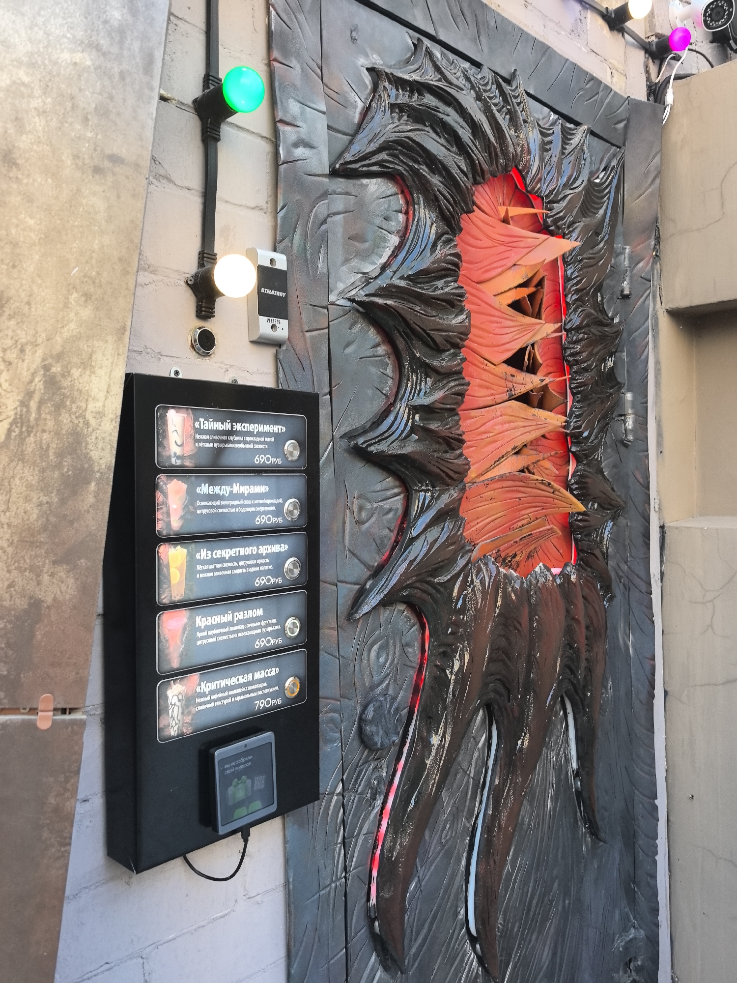 This image depicts an outdoor scene featuring an elaborate, theatrical doorway decorated to resemble a monstrous mouth or a portal, with jagged, dark-colored elements and glowing red and orange interiors. Adjacent to this doorway is a black box displaying a menu of drinks with their names, descriptions, and prices in Russian. The menu lists "Тайный эксперимент", "Между-Мирами", "Из секретного архива", "Красный разлом", and "Критическая масса", with prices in rubles. Below the menu is a small digital screen displaying a message in Russian that translates to "we have not forgotten your gift".
To the left of the menu box, a brick wall is visible, with a light fixture featuring a green bulb and another with a yellow bulb. A white security camera is mounted on a pole above the doorway, with a pink light attached to it. The environment appears to be an urban setting, possibly an entertainment venue or an escape room. Based on the presence of Cyrillic script and the specific currency, it is likely located in Russia. While the image itself does not definitively confirm Nizhny Novgorod, the style and context are consistent with attractions found in Russian cities.