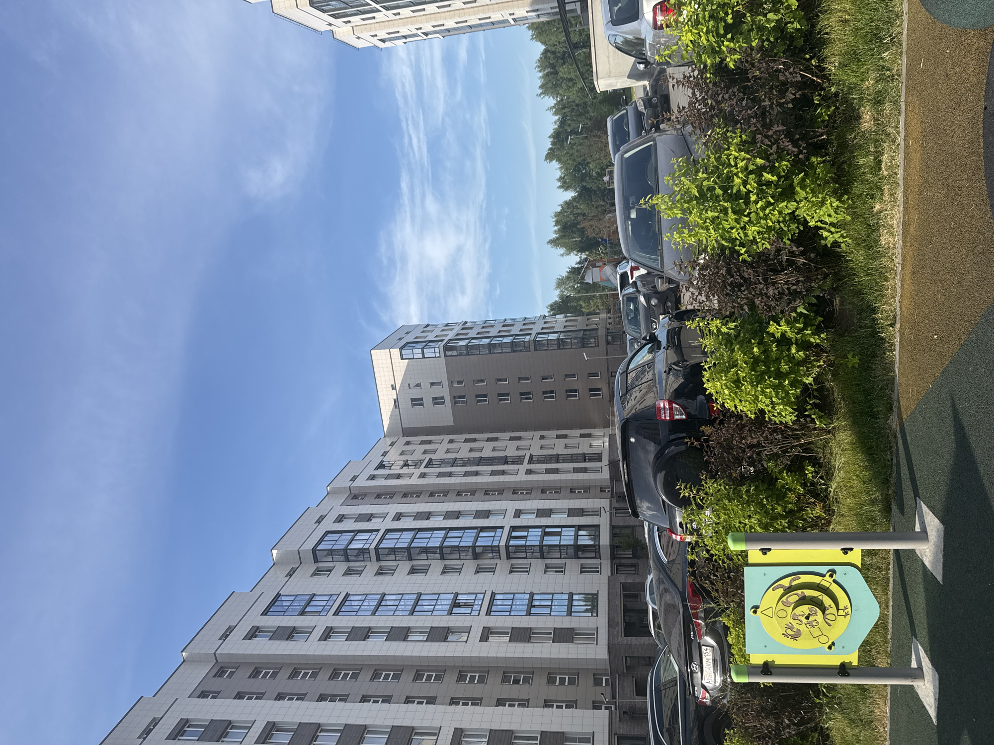 The image shows a modern residential complex with multi-story buildings. There are several cars parked in front of the buildings, including SUVs and sedans. Lush green bushes and trees surround the area. In the foreground, there is a children's playground equipment with colorful panels and posts. The sky is blue with scattered clouds. The scene appears to be in Koltsovo, Russia, given the context. There are no people visible in the image.