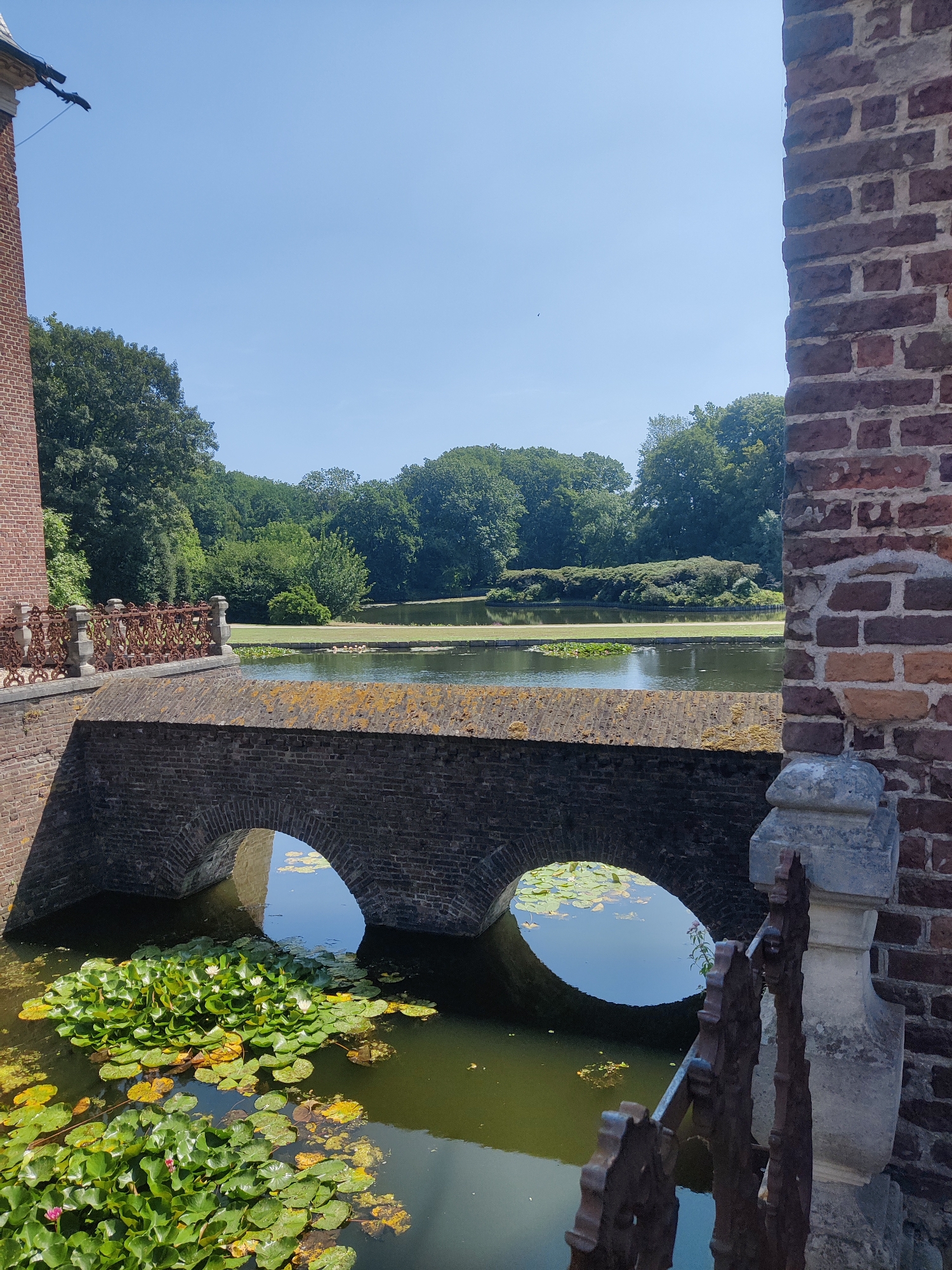 The image captures a view of the Isselburg Castle in Isselburg, Germany, on a sunny day. The foreground features a brick bridge with two arched openings over a body of water. Water lilies are abundant in the water below the bridge. To the left, a portion of a red brick tower with gargoyles is visible. On the right, more red brickwork forms part of the castle structure, with a decorative stone element and a rusted metal railing. Beyond the bridge, a tranquil lake is surrounded by lush green trees under a clear blue sky. There are no people or animals visible in the scene.