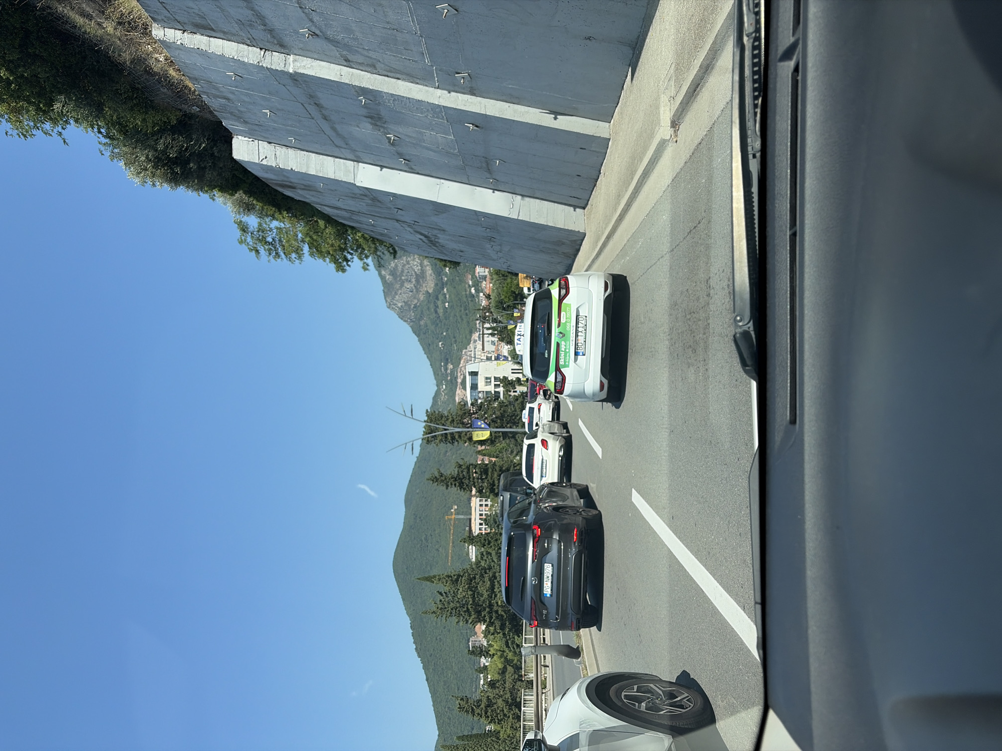 The image depicts a street scene in Budva, Montenegro, likely during the daytime on a clear, sunny day. The perspective is from inside a vehicle, looking out at a line of cars on a road.
The primary objects are several vehicles, including a white taxi with green lettering and a license plate reading "BO-TA 470". Behind it are other cars, including a dark gray SUV with the license plate "PG-AM 975". Further down the road, more vehicles are visible in traffic.
The setting includes a large, reinforced concrete wall on the left side of the road, which appears to be part of a hillside retaining structure. Lush green trees and vegetation are visible on the hillside above the wall and also lining the side of the road. In the background, a town with buildings and a prominent mountainous landscape are present. The sky is a clear, bright blue.
The activity appears to be a traffic jam, with cars stopped or moving slowly. There are no visible people in the scene. The overall mood is calm and ordinary, with no strong emotional cues.
Visible text includes "TAXI" on the white car, along with what appears to be a company name or app name, and a phone number "068 10 1577". The license plate on the dark gray SUV is also visible.