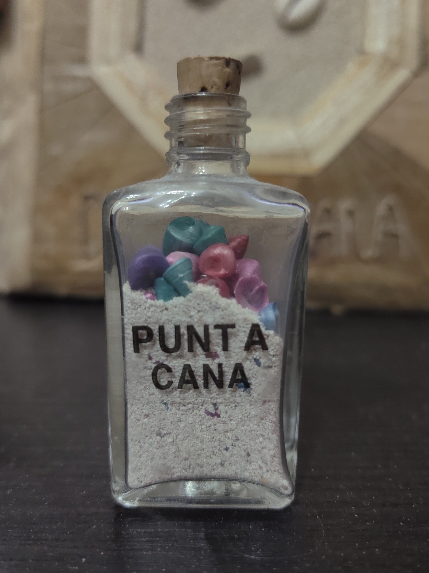 A clear glass bottle with a cork stopper stands on a dark, textured surface. The bottle contains light-colored sand, with small, colorful shell-like decorations mixed within the sand at the top. The words "PUNTA" and "CANA" are printed in black on the front of the bottle, over the sand. The background is blurred and appears to be a light-colored, textured object. The scene is well-lit, with the bottle being the primary subject. The location is not discernible from the image.