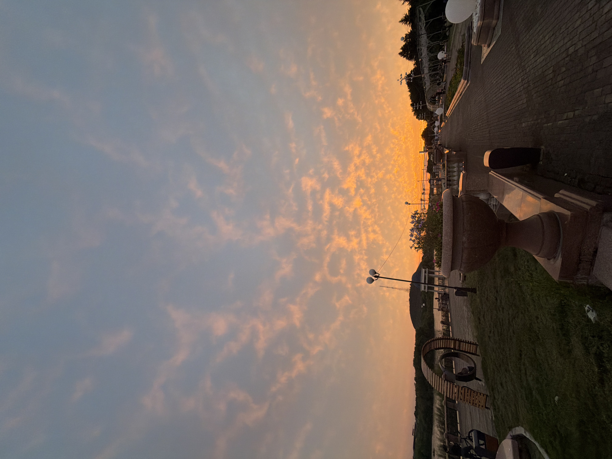 The image depicts a sunset scene at a riverside park. The sky is filled with scattered clouds tinged with orange and pink hues. A paved walkway extends into the distance, lined with decorative planters and lighting fixtures. On the left side of the walkway, a grassy area features a unique wooden sculpture and a body of water. In the background, a hill with a radio tower is visible. The park appears to be in a city environment, with trees and some buildings visible on the horizon. There are a few people scattered throughout the park, some walking and others engaged in activities near the sculpture. The overall impression is one of a tranquil evening in an urban park.