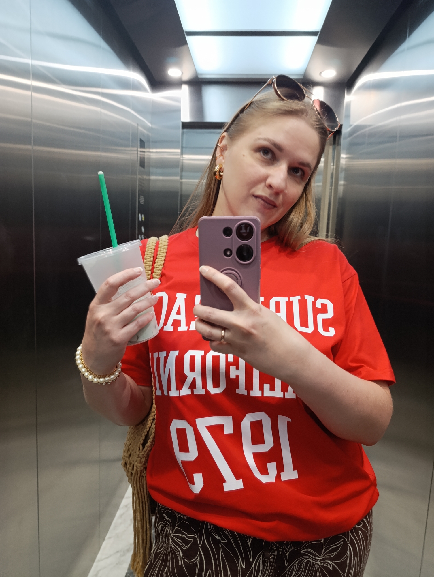 A woman stands in an elevator and takes a selfie. She has blonde hair and is wearing a red t-shirt with white lettering, sunglasses pushed up on her head, hoop earrings, a pearl bracelet, and patterned pants. She is holding a clear plastic cup with a green straw in her left hand and her purple phone in her right hand. The elevator walls are made of brushed metal and the ceiling has fluorescent lighting.