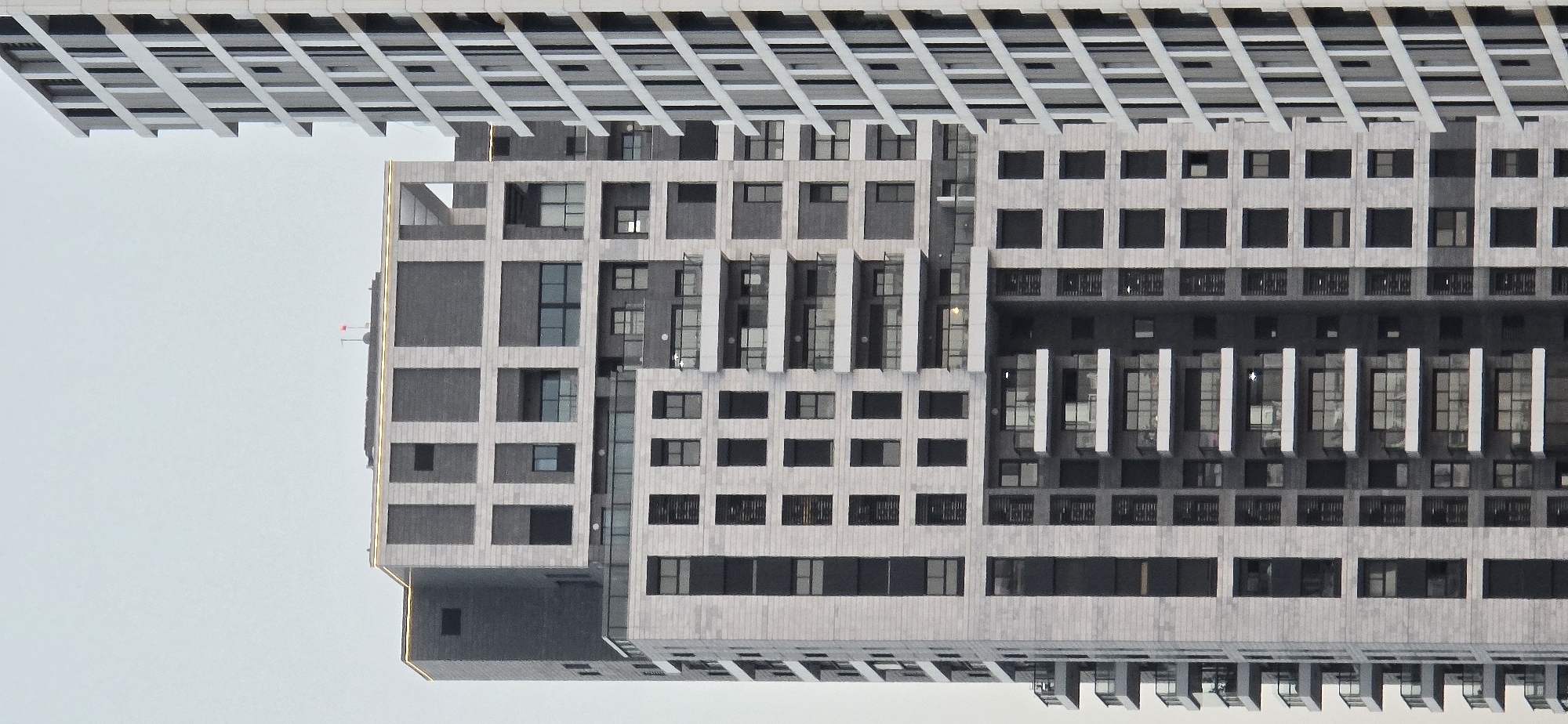 The image displays the facade of a modern, multi-story residential building, likely part of a complex in Bat Yam, Israel. The building features a distinctive architectural style with alternating light-colored stone and darker gray panels, arranged in a grid pattern. Many windows and balconies are visible, some with glass railings. To the right of the main building, another contemporary building with a linear, white facade is partially visible. The sky above is overcast and a pale gray. No people or other significant objects are discernible in the frame.