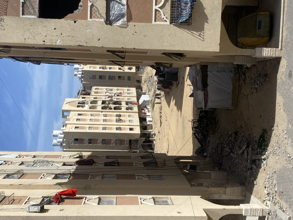 The image presents an urban scene in Khan Yunis, Palestinian Territory, characterized by residential buildings flanking an open, unpaved area under a clear blue sky with scattered clouds.
Two multi-story, light beige residential buildings frame the foreground. The building on the left exhibits multiple windows, some with clotheslines holding draped fabric, including a prominent red garment. Exposed electrical conduits are visible on its facade. The building on the right displays significant structural damage, notably a large, irregular, dark void on an upper-middle floor, and several windows covered with plastic sheeting or tarpaulin. Its ground level features an arched entrance containing a large, tarp-covered object, yellow-green in color, possibly a vehicle or equipment. Both foreground buildings have debris and rubble accumulated at their bases.
The central ground is a sandy, unpaved expanse heavily littered with scattered concrete rubble, refuse, and dark debris. In the mid-ground, a linear arrangement of approximately six makeshift shelters, constructed from various tarpaulins and materials, stretches horizontally. One shelter, specifically white, features a visible UNICEF logo. Adjacent to the base of the foreground right building, a distinct pile of charred wreckage and debris is present.
A small group of approximately five to seven individuals, appearing to be adults and children, traverse the sandy ground in the mid-distance, walking from left to right near the makeshift shelters.
In the background, several more multi-story residential buildings, similar in architectural style to those in the foreground, are visible. Many of these background structures feature multiple white cylindrical water tanks on their rooftops. Some also exhibit signs of damage or improvised coverings.