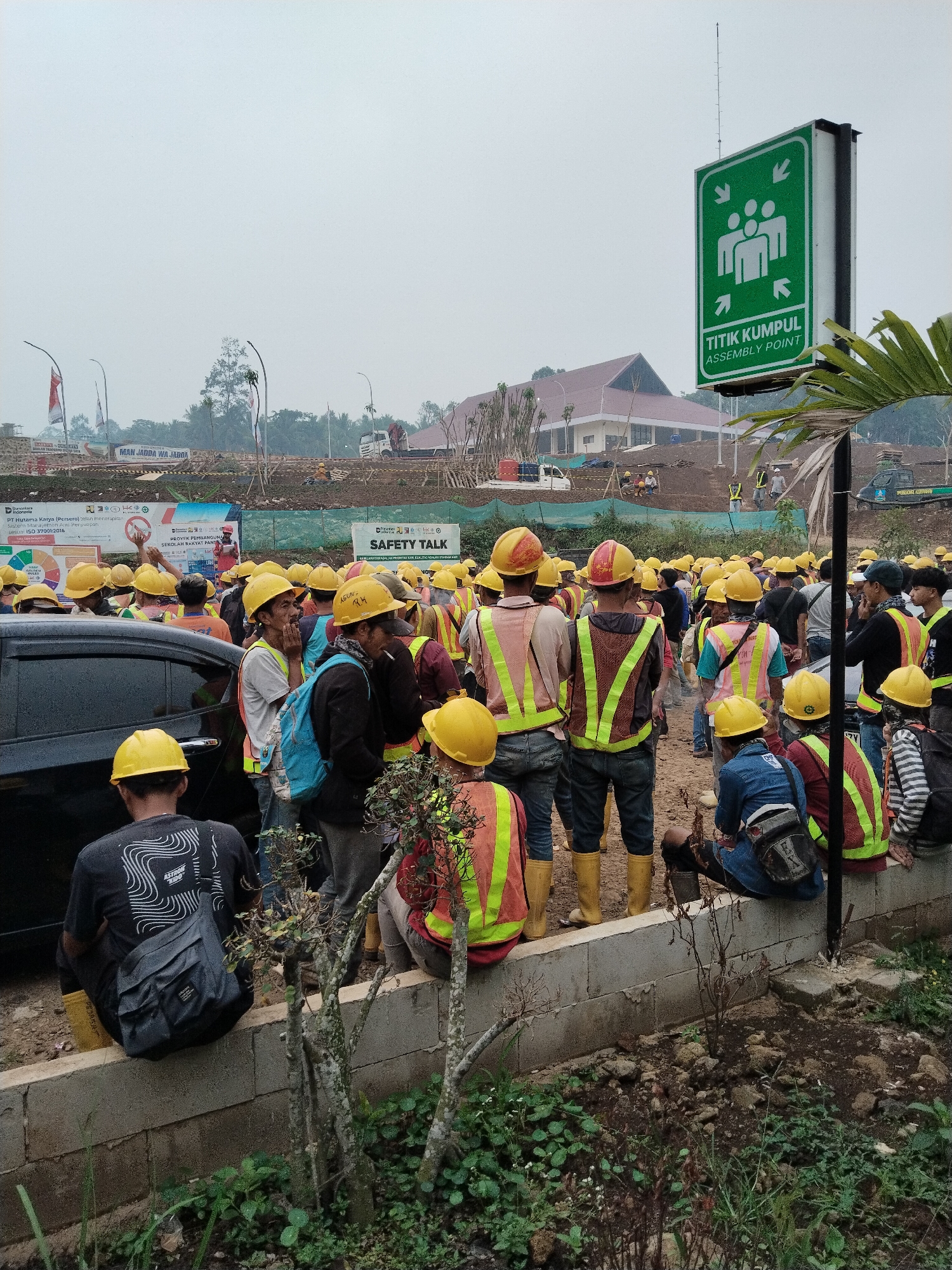 The image depicts a large group of construction workers gathered outdoors in what appears to be an industrial or construction site in Cemplang, Indonesia. The workers are predominantly wearing yellow hard hats and high-visibility vests in various colors, including yellow, orange, and green. Many are also wearing work boots, some of which are yellow. The group is assembled near a wall, with some individuals standing and others sitting on the wall.
In the background, a large building with a brown roof is visible, along with construction equipment and materials. There is also a green sign with white iconography and text that reads "TITIK KUMPUL ASSEMBLY POINT," indicating a designated gathering area. A black car is parked on the left side of the image. The overall environment suggests a work-in-progress setting, with signs of ongoing construction activities. The sky is overcast, indicating a cloudy or hazy day. A few trees and some vegetation are present in the foreground and background.