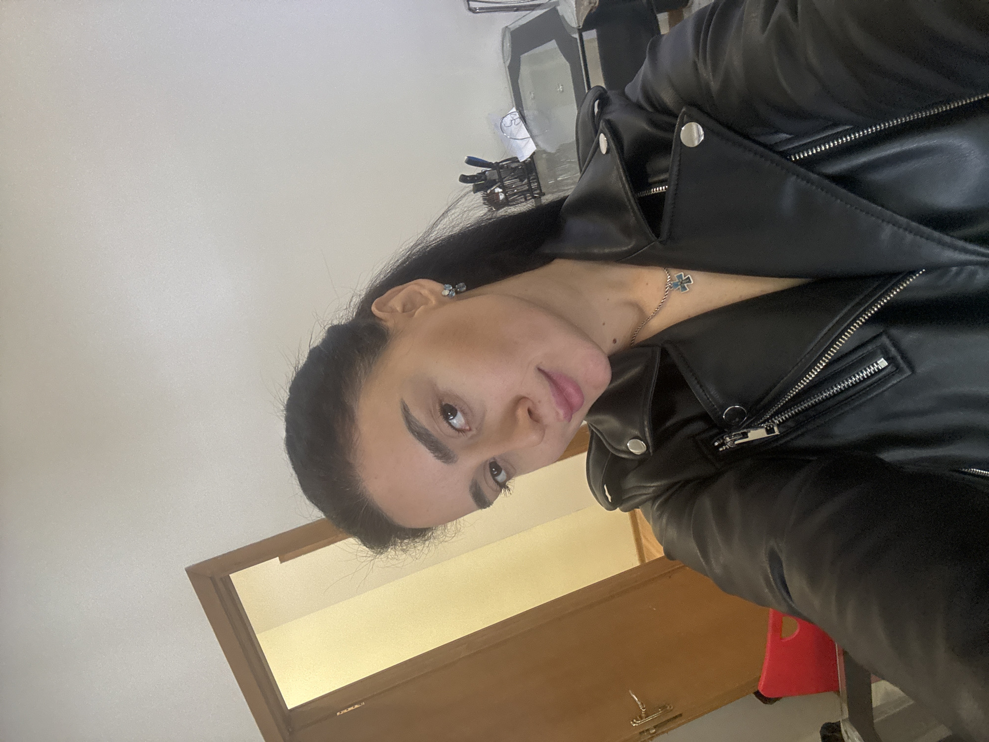A person with dark hair is wearing a black leather jacket. They are indoors, possibly in an office or apartment, as suggested by the door in the background. The presence of what appears to be a rolling office chair and various items on a desk (partially visible in the background) further supports this. The person is also wearing a necklace. The location is impossible to determine with the given information.