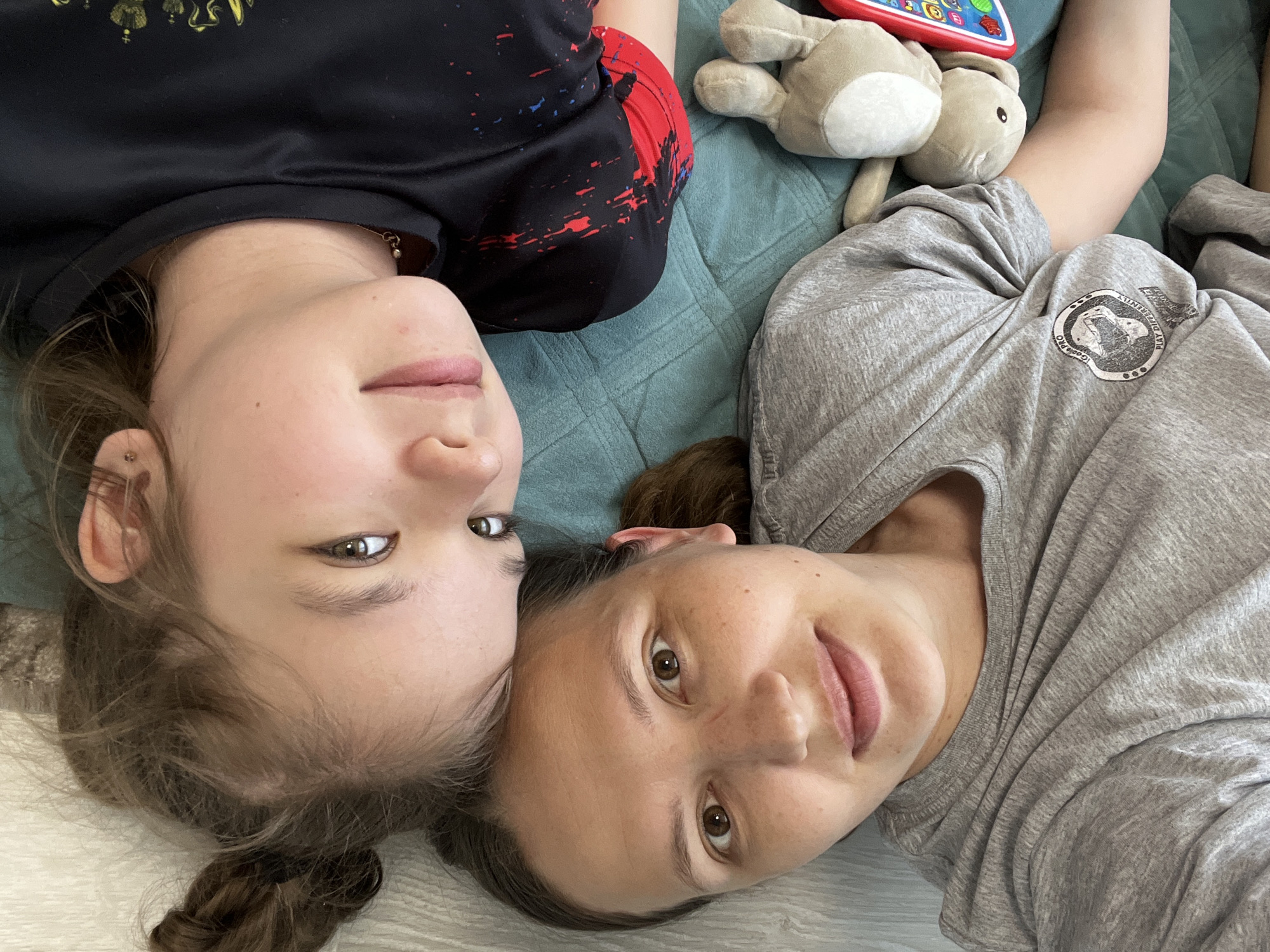 Two people, likely a mother and daughter, are lying on a blue-green quilted bedspread. The location is suspected to be in Chaikovsky, Russia. The person on the left is wearing a black shirt with red and blue designs. The person on the right is wearing a grey t-shirt. Next to the two people is a grey stuffed animal rabbit and a children's toy with colorful buttons. The background consists of the bedspread, and part of a rug or carpet in the lower left corner.