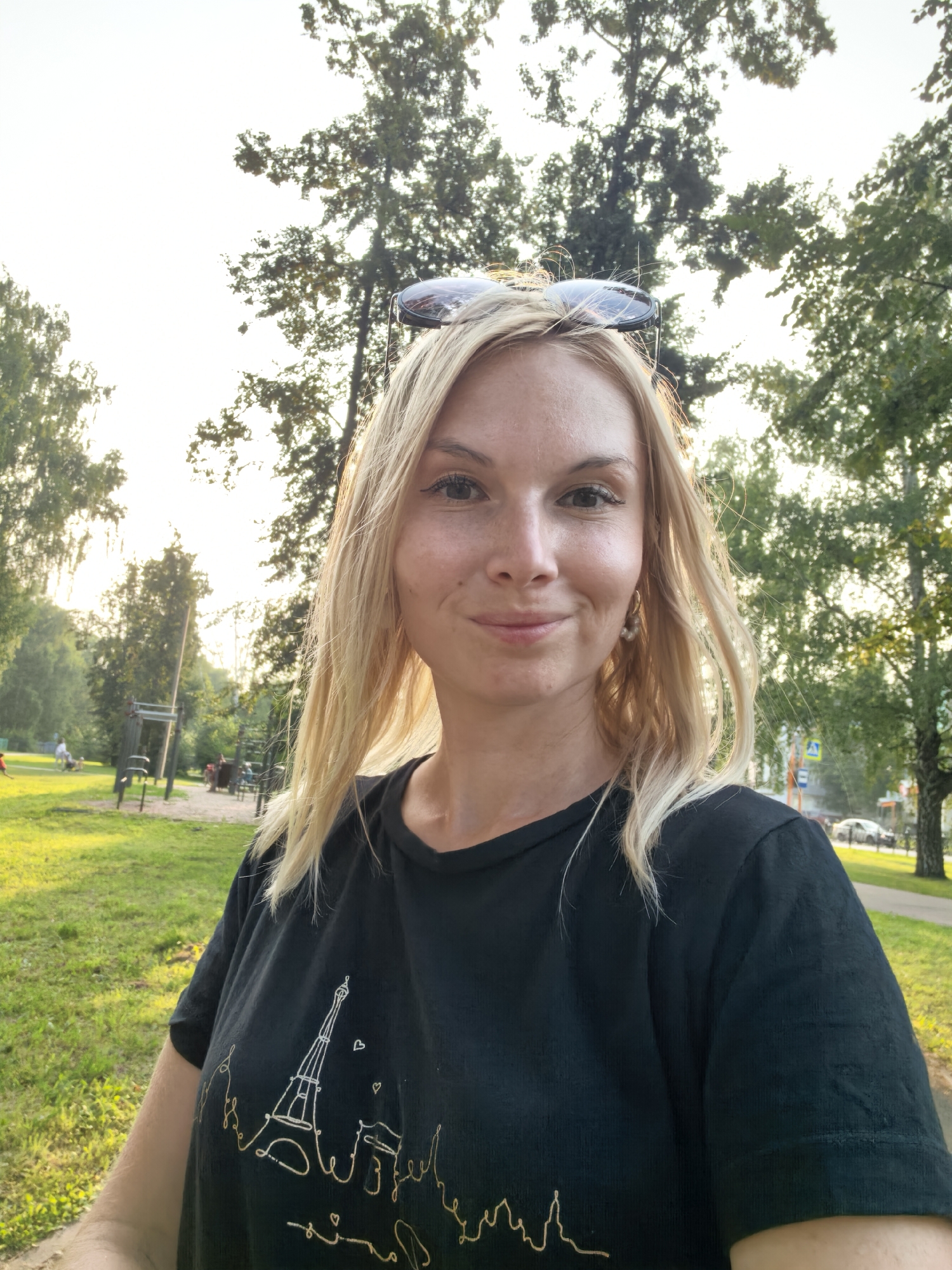 The image shows a young Caucasian woman with blonde hair and fair skin, wearing sunglasses on her head and a black t-shirt with an Eiffel Tower design. She is outdoors, with trees and a park-like setting behind her. The lighting suggests it is daytime, possibly late afternoon, with the sun filtering through the leaves. The environment includes green grass, trees, and a path or road in the background with some distant cars and buildings. There are also hints of outdoor recreational equipment in the middle ground. The overall impression is a casual portrait taken in a park or public outdoor space. The exact location in the city of Sterlitamak, Russia, cannot be determined from the visual information provided in the image.
