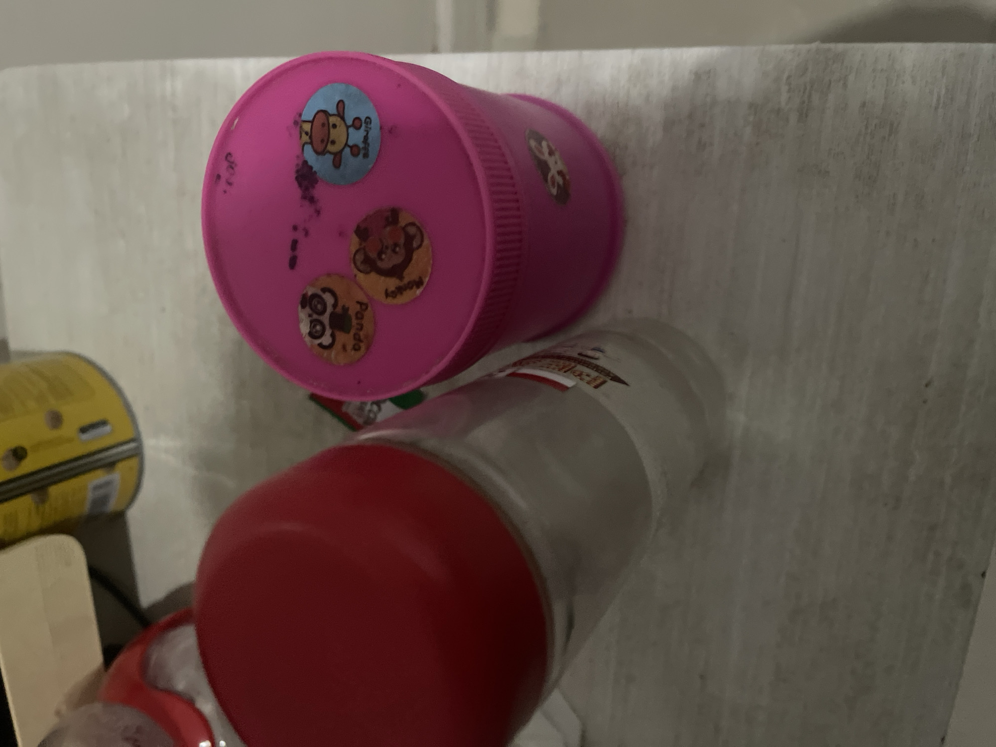 This image shows a close-up, angled view of a light-colored surface, possibly a wall or a shelf, with several objects placed on it. The primary focus is on a pink plastic cup or container, decorated with several round stickers featuring cartoon animals. One sticker depicts a giraffe, another a monkey, and a third appears to be a panda. Some of these stickers have text, with "Giraffe" clearly visible on one, and "Monkey" and "Panda" potentially visible on others. The cup itself has some dark smudges on its surface.
To the lower right of the pink cup, a clear glass jar with a red lid is visible. The jar contains some white material, possibly a powder or small objects. A label with red and white text is present on the upper portion of the glass jar, but the text is largely illegible due to the angle and lighting.
Further to the left, a yellow cylindrical object with black and yellow markings is partially visible. This object appears to be a container or can, possibly for food or another product. A thin, light-colored wooden object, resembling a ruler or a piece of trim, is positioned in the lower left corner of the frame, casting a shadow.
The scene is dimly lit, suggesting an indoor environment, and there are no clear indicators of time of day or weather. The overall impression is of everyday objects arranged casually, possibly in a home or storage area. There are no people visible in the image, and the setting is ambiguous.
