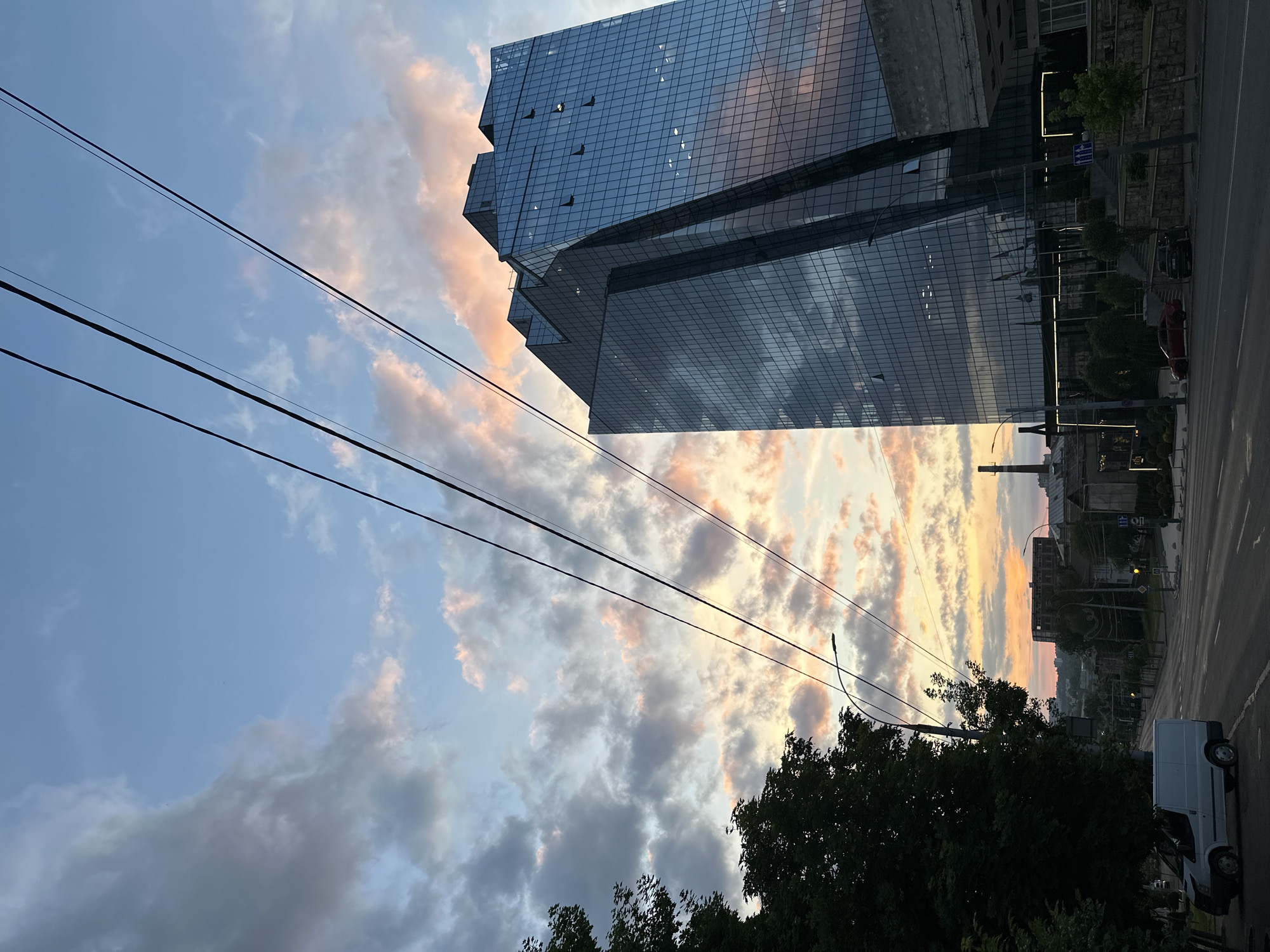 An angled, wide shot, taken in Chișinău, Moldova, depicts a modern glass-covered building under a sky with a striking sunset. The building reflects the sky. High-tension electrical wires stretch across the frame. The lower portion of the image is dominated by leafy trees. A white van is parked along the roadside. Another building can be seen, along with some infrastructure.