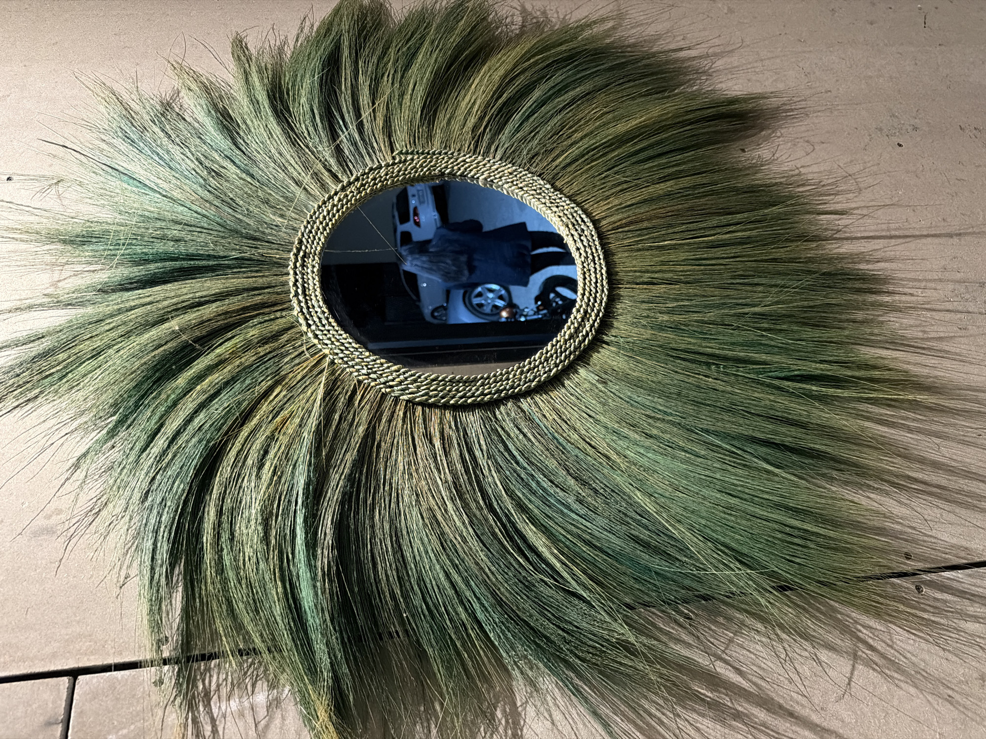 An oval-shaped mirror, framed by a circular arrangement of long, green, grass-like fibers that radiate outwards, is mounted on a taupe-colored wall. The fibers are a mixture of green and yellow hues and are densely packed around the mirror, creating a sunburst or feather-like effect. The wall has a textured surface and a slight sheen, with some visible lines that suggest paneling or tiles. In the reflection of the mirror, a woman with long dark hair is visible from the back, wearing a dark coat and dark leggings. She is standing in what appears to be a garage or driveway, with a white car and a motorcycle reflected behind her. The overall scene is an indoor shot, likely in a home or commercial space in Burgas, Bulgaria.