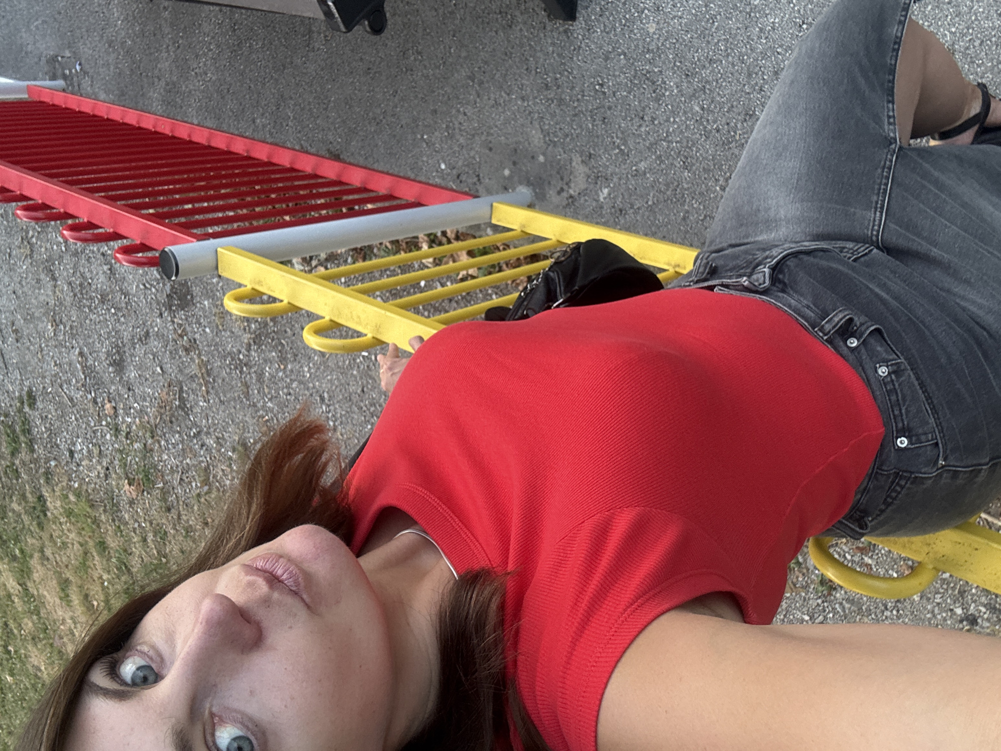 A woman with auburn hair and blue eyes is posing for a selfie outdoors. She is wearing a red short-sleeved shirt and dark gray denim shorts. A black purse is slung over her shoulder. She is leaning against a yellow railing, with a red fence in the background. The ground is covered in gravel and sparse grass. The lighting suggests it is daytime.