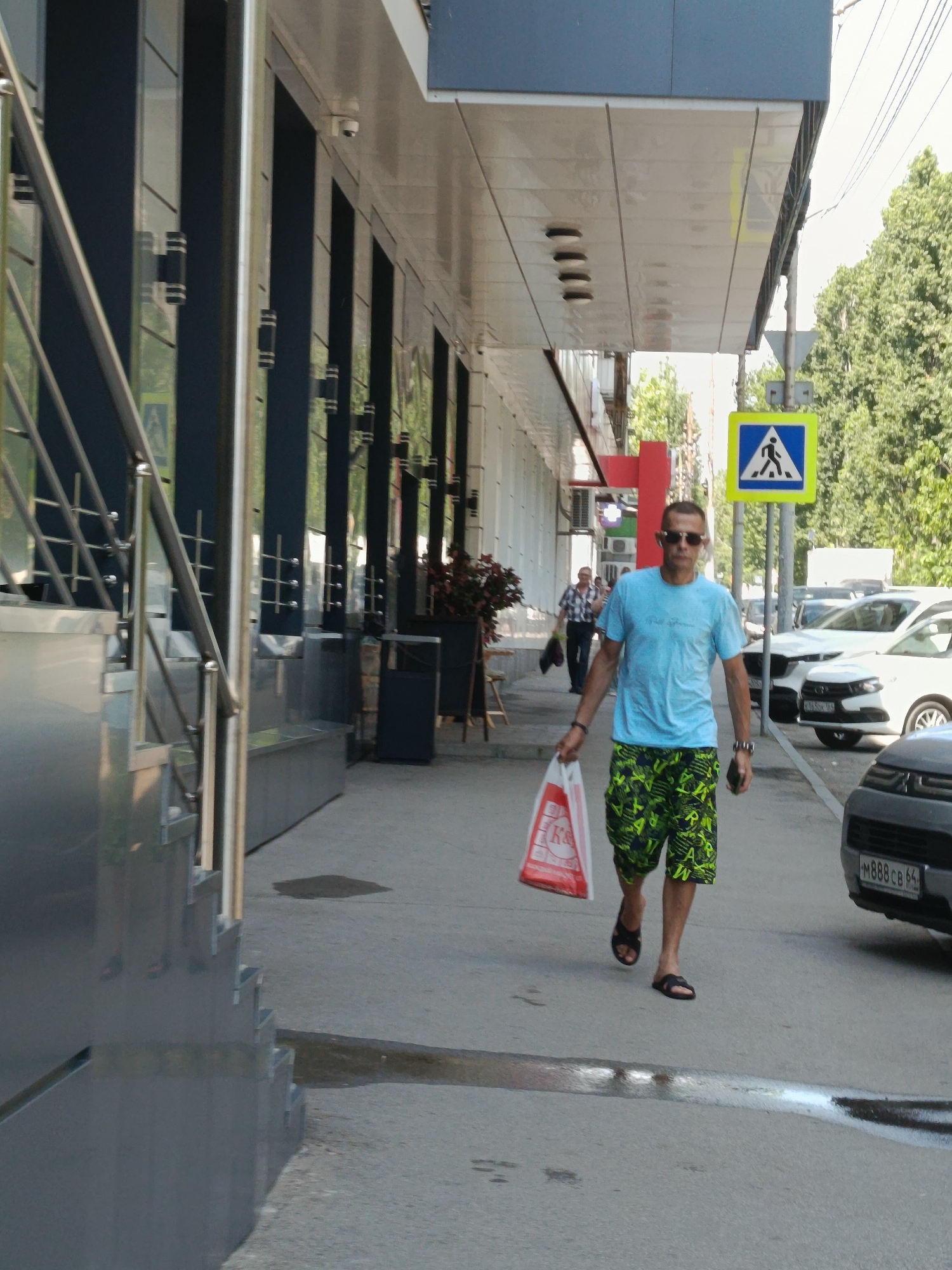 A man is walking down a sidewalk in Saratov, Russia, carrying a red and white plastic bag in his left hand. He is wearing a light blue t-shirt and green and black patterned shorts, along with black flip-flops. The man is wearing sunglasses. Behind him, a pedestrian crossing sign is visible on a pole. The sidewalk is paved with concrete, and there are trees and buildings in the background. A dark gray car is parked on the right side of the frame.