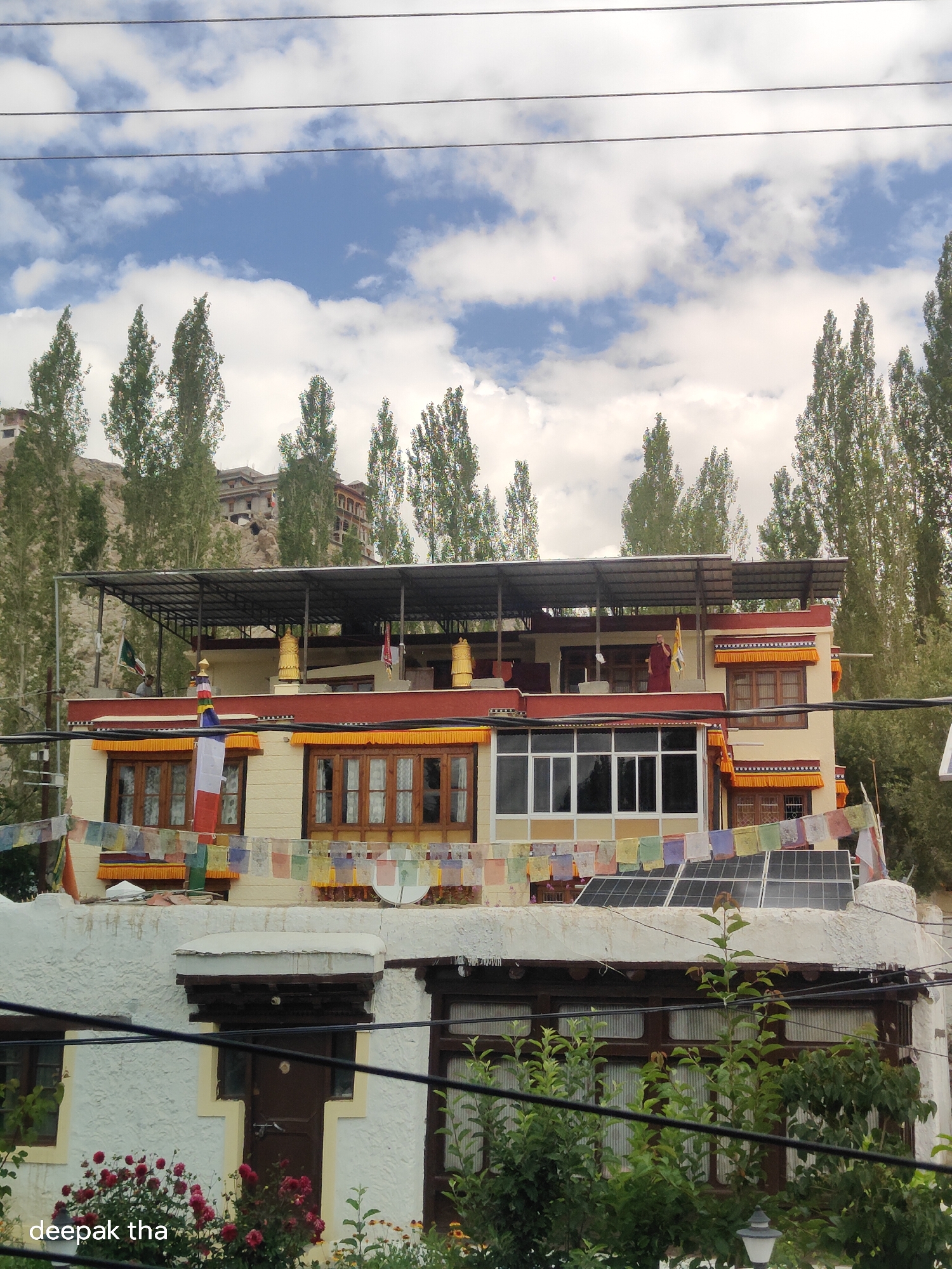 The image shows a multi-story building with a red-and-yellow color scheme, set against a backdrop of tall, slender trees and a mountainous landscape under a cloudy sky. Prayer flags are strung across the building, and solar panels are visible on the roof. A person in a maroon robe stands on the upper level of the building. In the foreground, there is dense foliage, red roses, and the text "deepak tha" overlaid on the bottom left corner. The scene suggests a location in Leh, India, possibly a monastery or a guesthouse, given the architectural style and the prayer flags. The overall atmosphere is serene and appears to be a daytime shot.