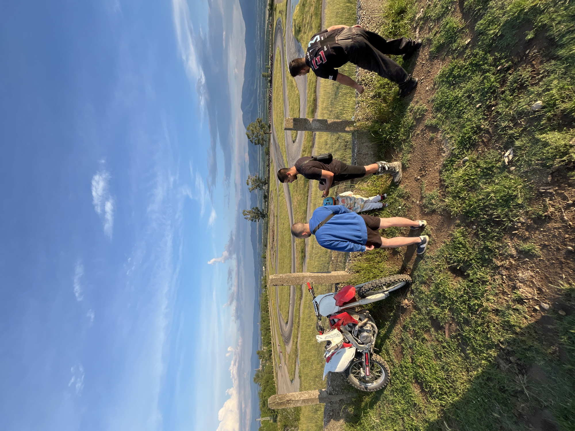 The image, correctly oriented, depicts an outdoor scene at what appears to be a motocross track, set against a mountainous backdrop near Anevo, Bulgaria. The time of day is late afternoon or early evening, characterized by warm, golden light and long shadows, under a clear blue sky with scattered white clouds.
The perspective is from a grassy, elevated bank, overlooking a winding dirt track that stretches through green fields and sparse trees in the mid-ground. Distant mountains frame the horizon. In the immediate foreground, three adult males and a small child are present. One man, wearing a black t-shirt with white markings (possibly 'M1' or 'H1') and black pants, leans forward, observing the track below. Another man in a black t-shirt and shorts, with a black shoulder bag, is positioned similarly, also looking towards the track. A third man, dressed in a blue long-sleeved top and black shorts, stands near a red and white dirt bike. The motorcycle has visible branding, including "BREMBO" and "MOTUL" on its tires, and is parked on the grassy terrain. A small child, likely a toddler or infant, lies on the ground between two of the men, wearing light-colored clothing and a light blue helmet. Two concrete posts are also visible in the foreground, bordering the edge of the elevated area. The adults appear to be observing the track, potentially in preparation for or watching motocross activities.