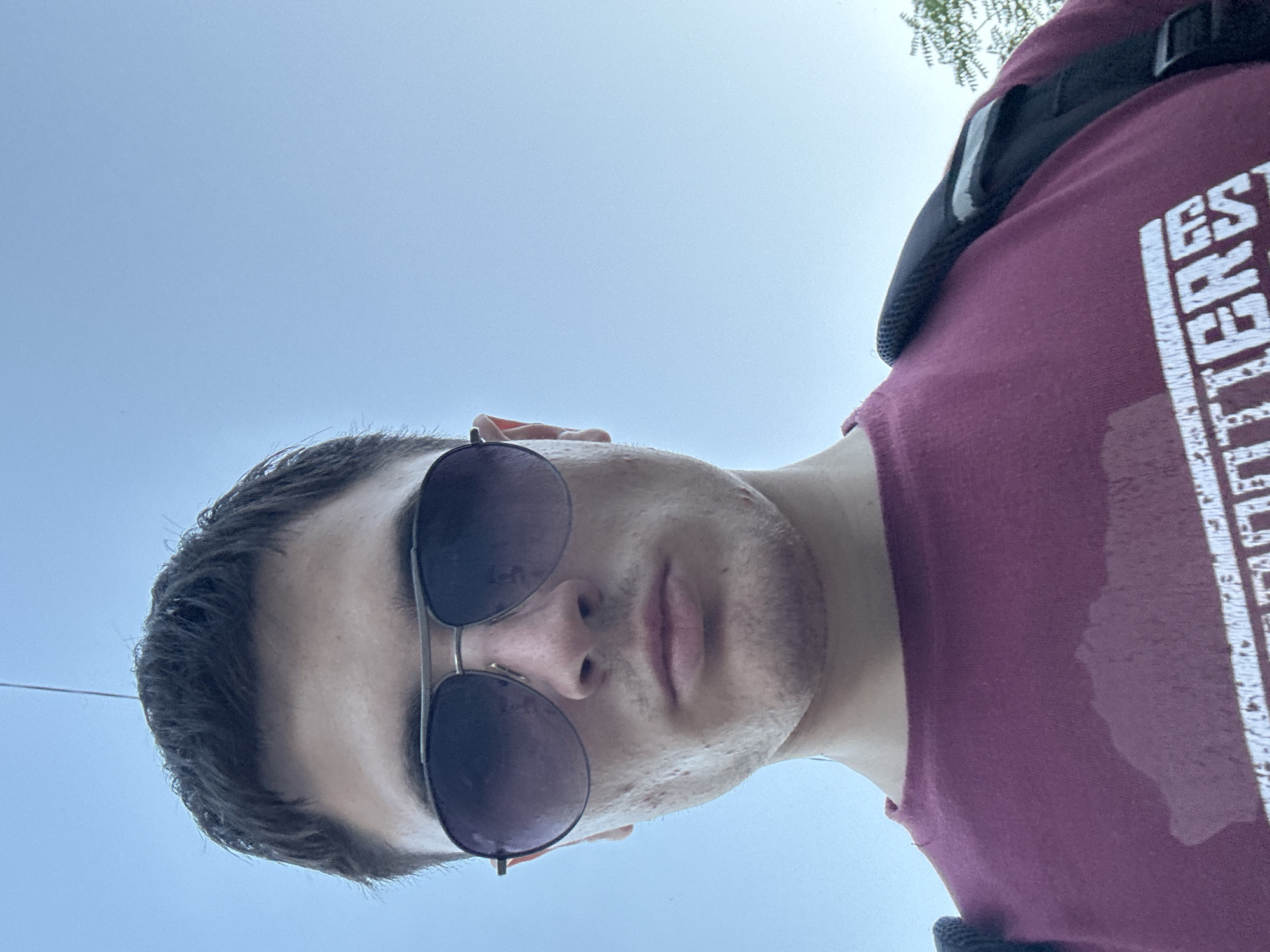 A young adult male, wearing aviator sunglasses and a maroon t-shirt, is captured from a low-angle perspective, looking towards the camera. The t-shirt exhibits noticeable sweat stains on the chest area, indicating a warm outdoor environment. The man's face is partially obscured by the sunglasses, but his expression appears serious. He is wearing a black backpack strap across his right shoulder. The background consists of a clear, light blue sky with a faint vertical line suggesting an overhead wire or pole. The setting is likely an outdoor public space in Stara Zagora, Bulgaria, given the context provided.