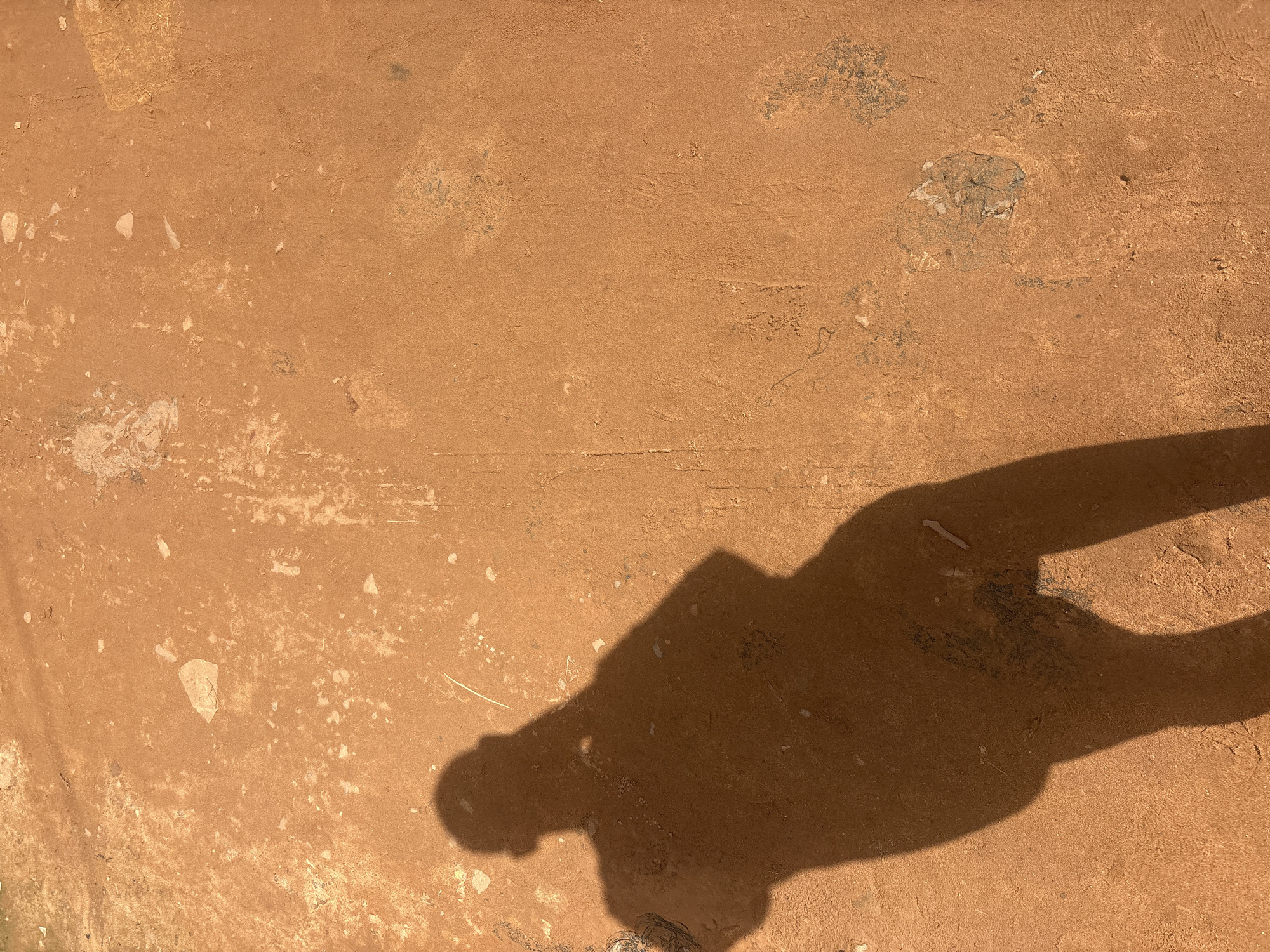 A shadow of a person is cast on a dusty, orange-brown surface in Nkpor, Nigeria. The shadow is in the lower left portion of the image and appears to be of someone holding a phone or camera to take a picture. The surface is uneven and shows signs of wear and tear, with small rocks and patches of lighter-colored material scattered across it. The light suggests it is daytime, and the shadow is sharp, indicating direct sunlight.