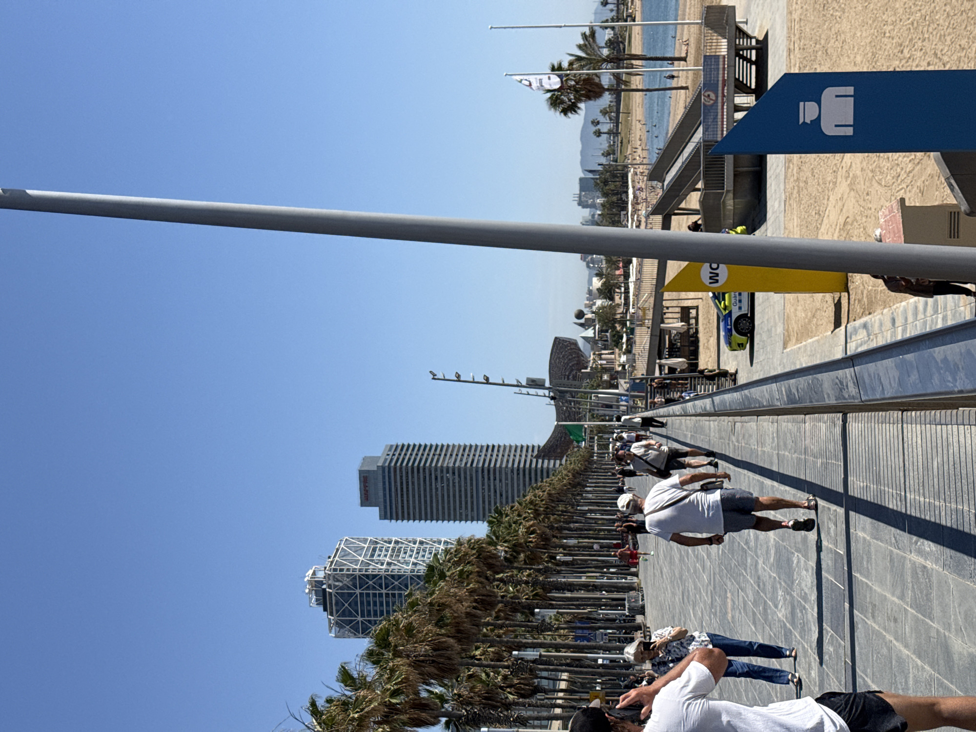 The image shows a sunny day at the Barceloneta beach in Barcelona, Spain, with the iconic Hotel Arts and Mapfre Tower in the background. A paved promenade runs along the beach, lined with palm trees. Several people are walking on the promenade, some casually strolling and others appearing to exercise. A police car is parked near a sign indicating public restrooms. The beach itself is visible, with people sunbathing and swimming in the Mediterranean Sea. The overall scene depicts a vibrant and active beach environment in a major European city.