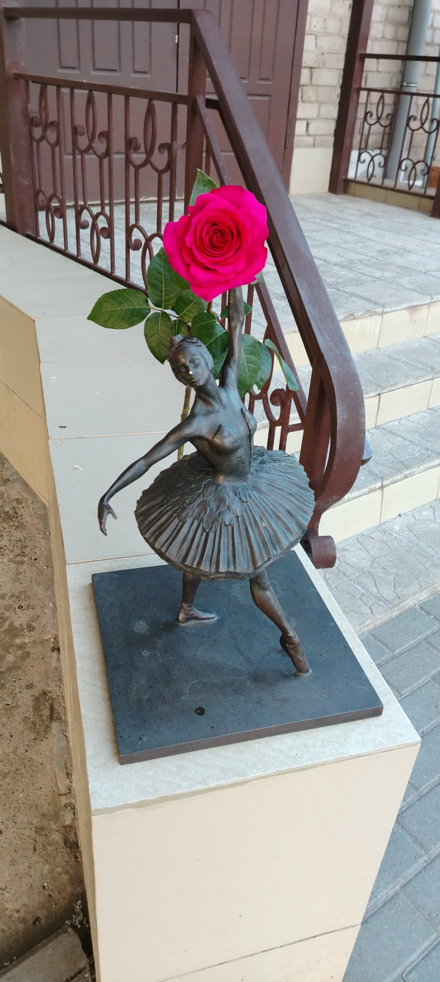 A bronze statue of a ballerina, holding a bright pink rose, stands on a beige pedestal beside a brown metal handrail in Chaikovsky, Russia. The ballerina is in a classical pose, wearing a tutu and ballet shoes. The base of the statue is a dark grey rectangle. The pedestal is made of beige stone and sits on a paved surface of grey and white tiles. Behind the handrail, a set of stone steps can be seen.
