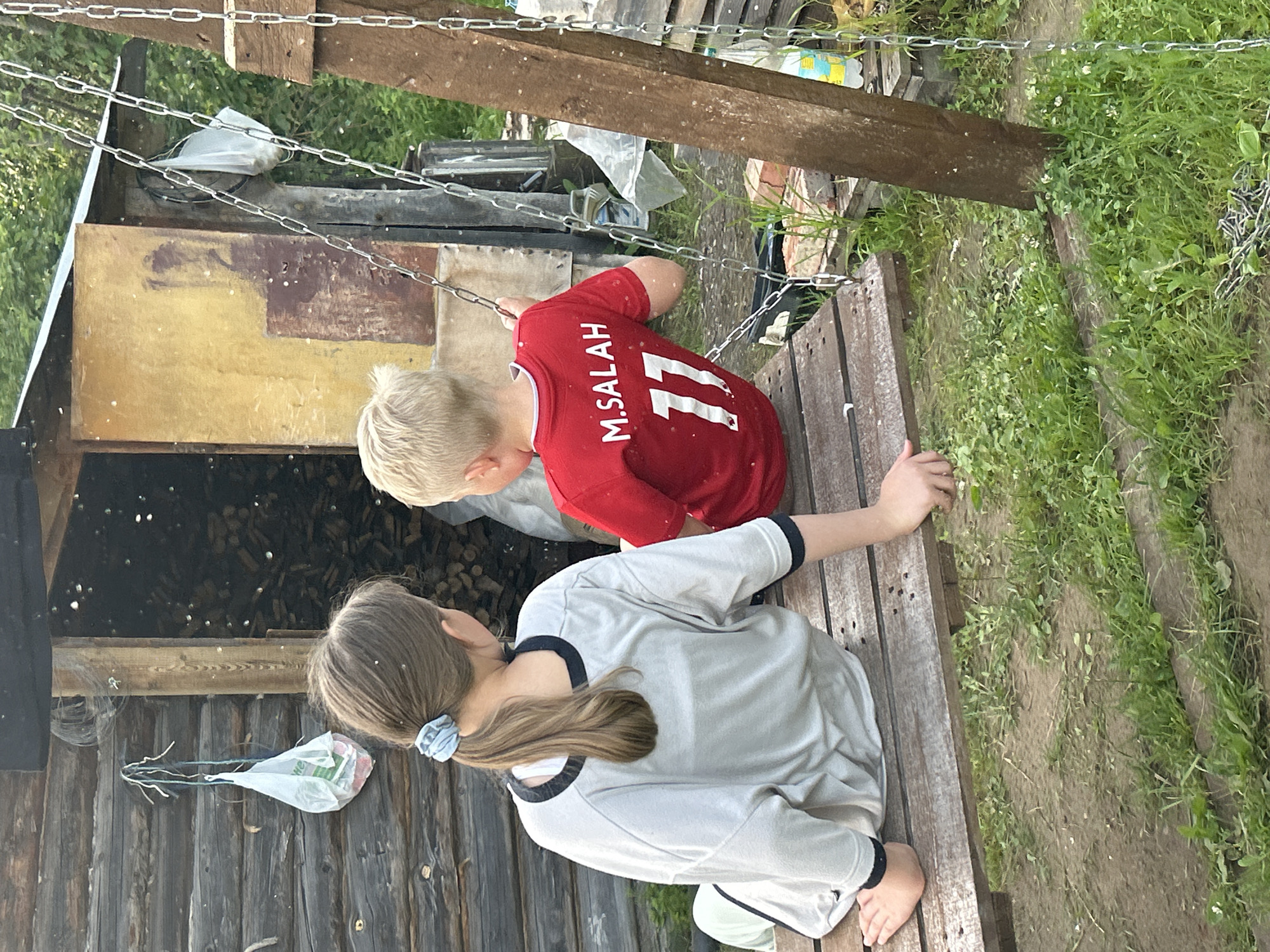 Two children, a girl and a boy, sit on a wooden swing set. The girl, on the left, has her back to the camera, with her brown hair tied in a ponytail. She wears a light-colored t-shirt with dark trim and light-colored pants. The boy, on the right, also has his back to the camera, wearing a red t-shirt with "M.SALAH 11" printed on the back. They are outdoors, with a rustic wooden building and stacked firewood visible behind them. The ground is covered in grass and dirt. This scene is located in the city of Chaikovsky, Russia, a city known for its industrial landscape and proximity to the Kama River. The image captures a moment of leisure in what appears to be a rural or semi-rural setting within the city.